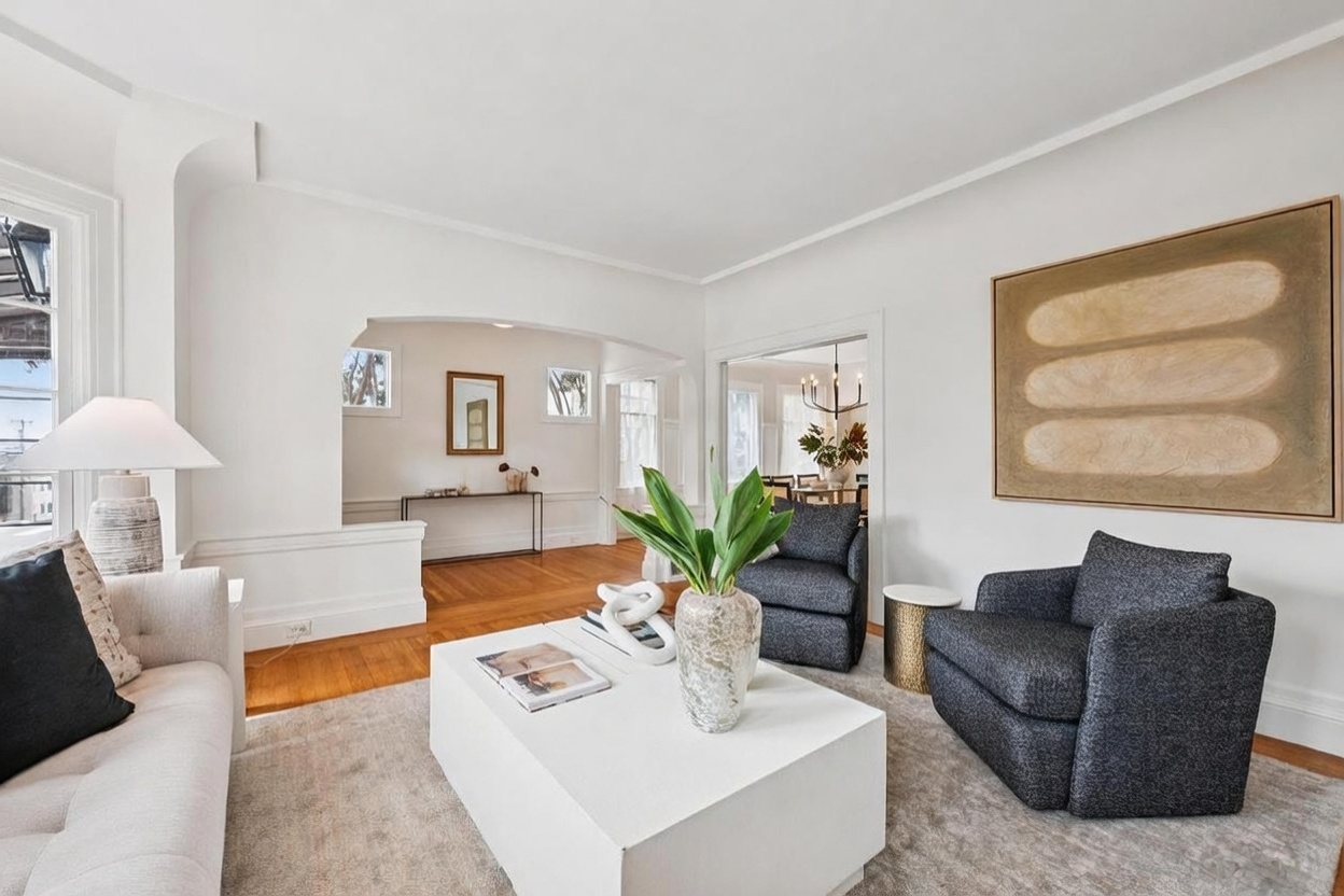 This bright and airy living room features a neutral color palette, plush seating, and a large, minimalist white coffee table as a central focal point. The space is accented by a large abstract art piece on the wall and flows seamlessly into an adjacent dining area through a wide, arched opening. The room is bathed in natural light, creating a sophisticated and welcoming atmosphere suitable for modern living.