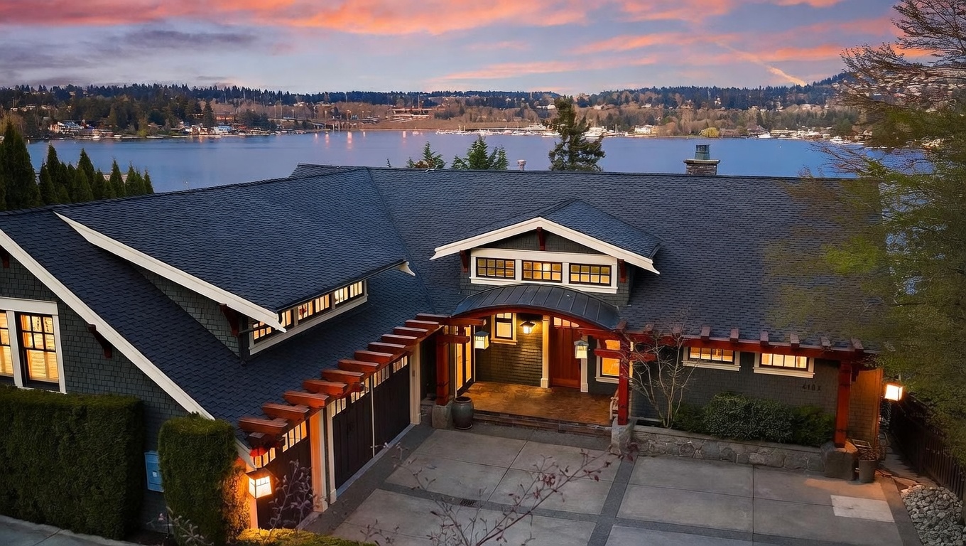 This high-angle exterior shot captures a stunning craftsman-style home at dusk, featuring dark shingle siding, a prominent arched entryway, and a decorative wooden pergola over the garage. The property is situated directly on a waterfront, with a serene lake view visible in the background under a vibrant, colorful sunset sky. The scene conveys a sense of luxury and tranquility, highlighting the home's architectural character and prime location.