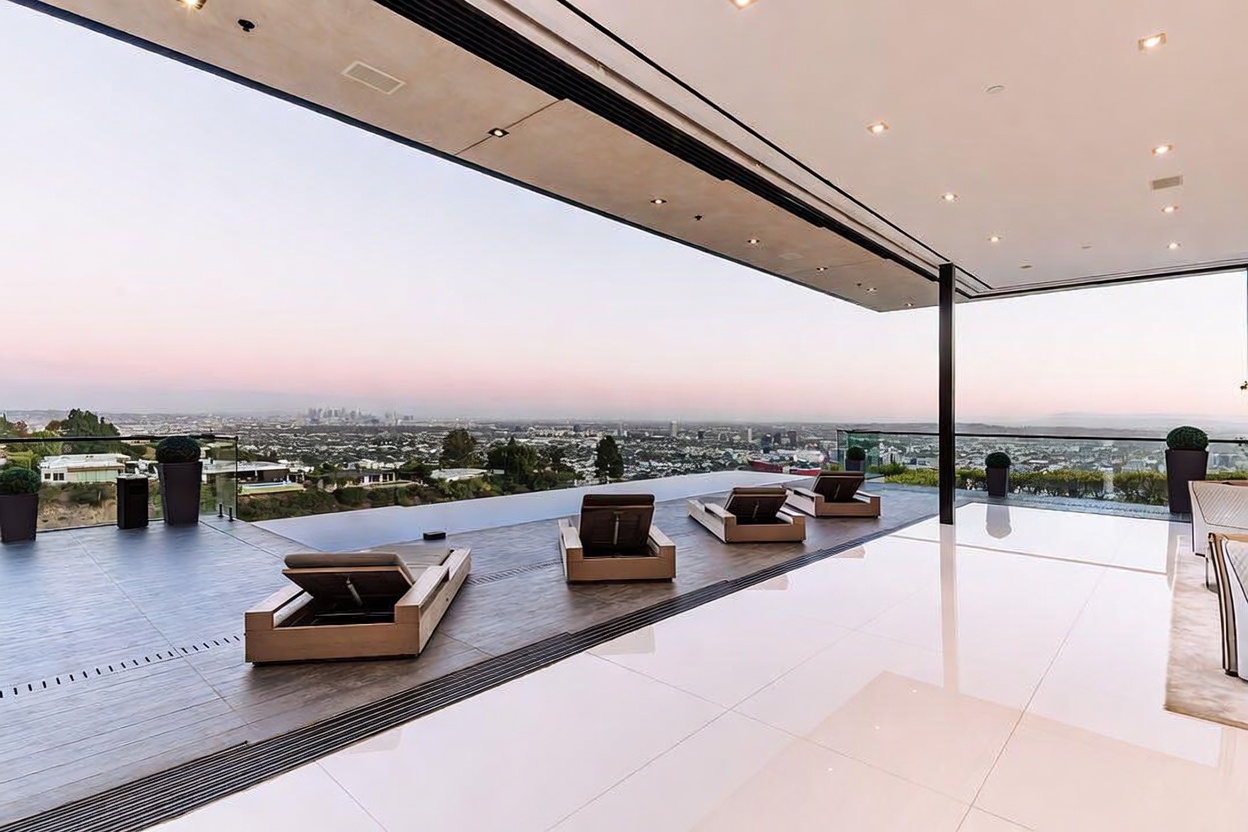 This luxurious indoor-outdoor living space features a seamless transition from a polished white interior floor to a dark-tiled deck overlooking an infinity pool and a sprawling city skyline. The area is furnished with modern lounge chairs, creating a sophisticated atmosphere perfect for relaxation and entertaining. The expansive, open-concept design emphasizes the panoramic views, highlighting the property's high-end architectural appeal.