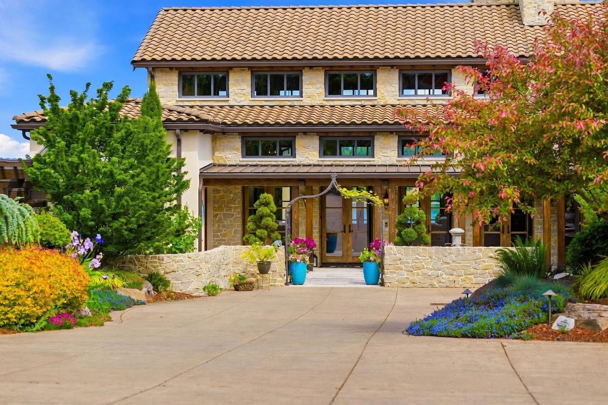 This front-facing view showcases a stately two-story home with a textured stone facade and a warm-toned tile roof. A paved driveway leads to a welcoming entryway framed by a decorative metal archway, stone retaining walls, and vibrant landscaping. The home features symmetrical window placement and a blend of rustic stone and wood accents, creating an inviting and upscale curb appeal.