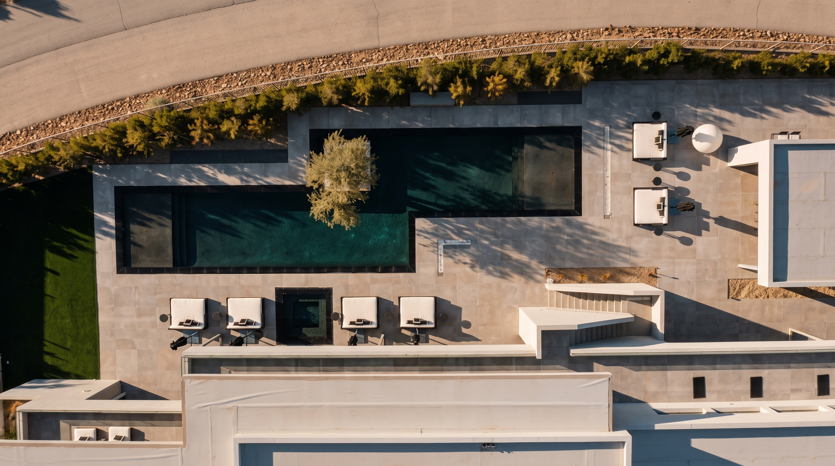 This high-angle aerial view captures a modern backyard oasis featuring a sleek, rectangular swimming pool with a central tree island. The surrounding patio is paved with large, light-toned stone tiles and is furnished with multiple lounge chairs and outdoor seating areas. The space is bordered by a manicured hedge and a retaining wall, creating a private and sophisticated outdoor living environment.
