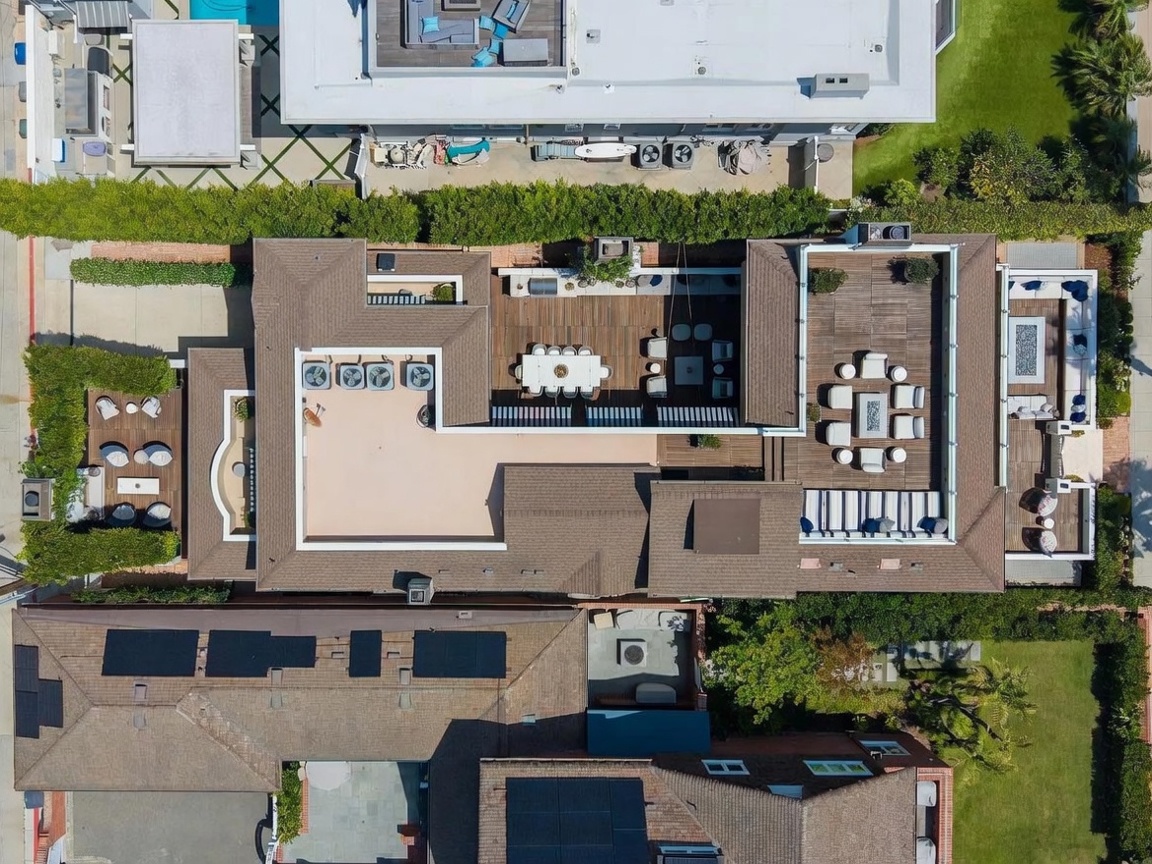 This high-angle aerial view captures the expansive rooftop layout of a multi-level residential property. The design features multiple interconnected wooden decks, including a central dining area, a lounge space with a fire pit, and various seating arrangements, all framed by lush green hedges and neighboring rooftops. The perspective highlights the sophisticated outdoor living spaces and the structured, layered architecture of the home.