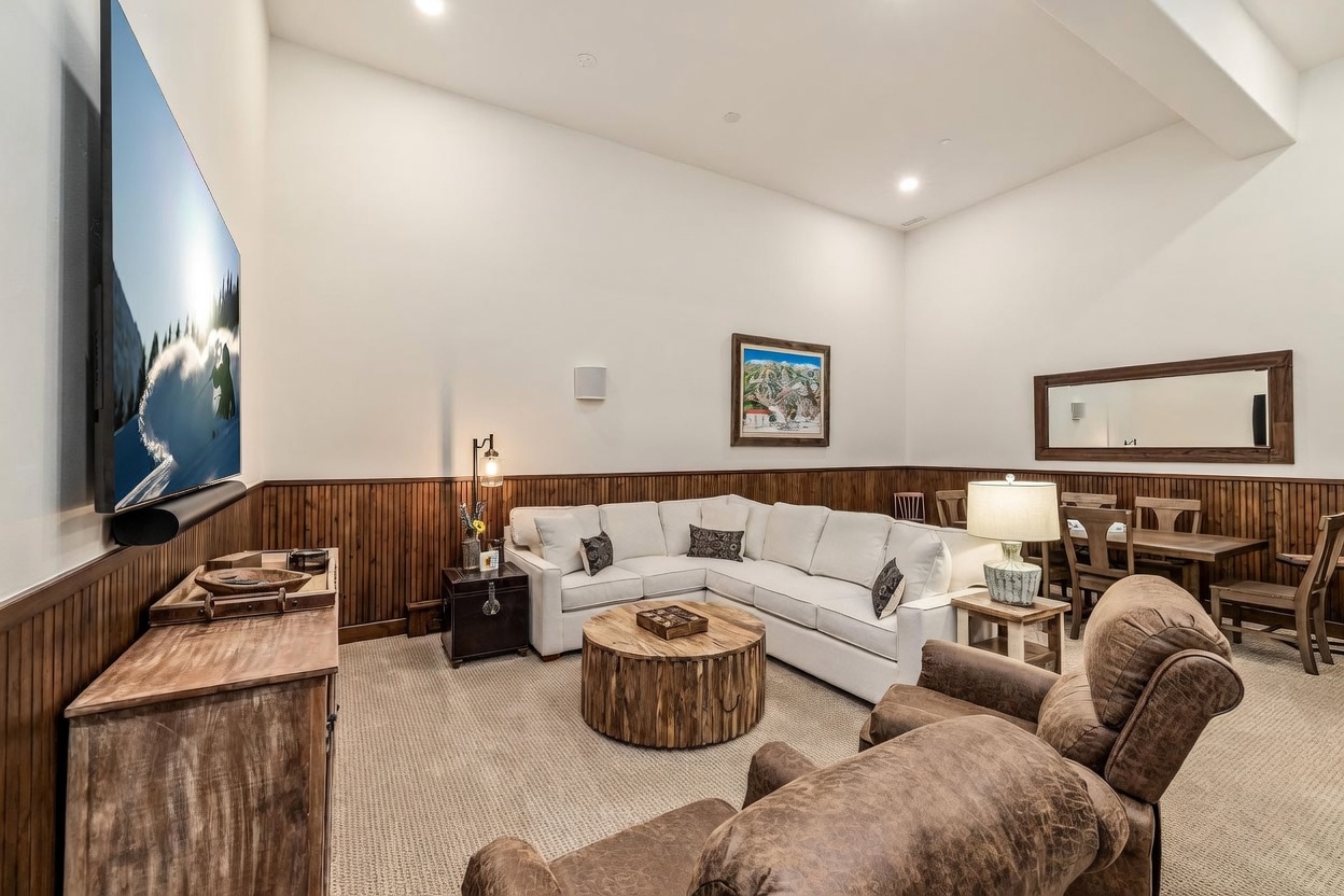 This spacious living room features a cozy, neutral-toned sectional sofa arranged around a rustic, cylindrical wood coffee table. The room is accented by wood wainscoting along the lower walls, a large wall-mounted television, and comfortable leather armchairs in the foreground. A dining area is visible in the background, creating an open-concept feel that is perfect for entertaining and relaxation.