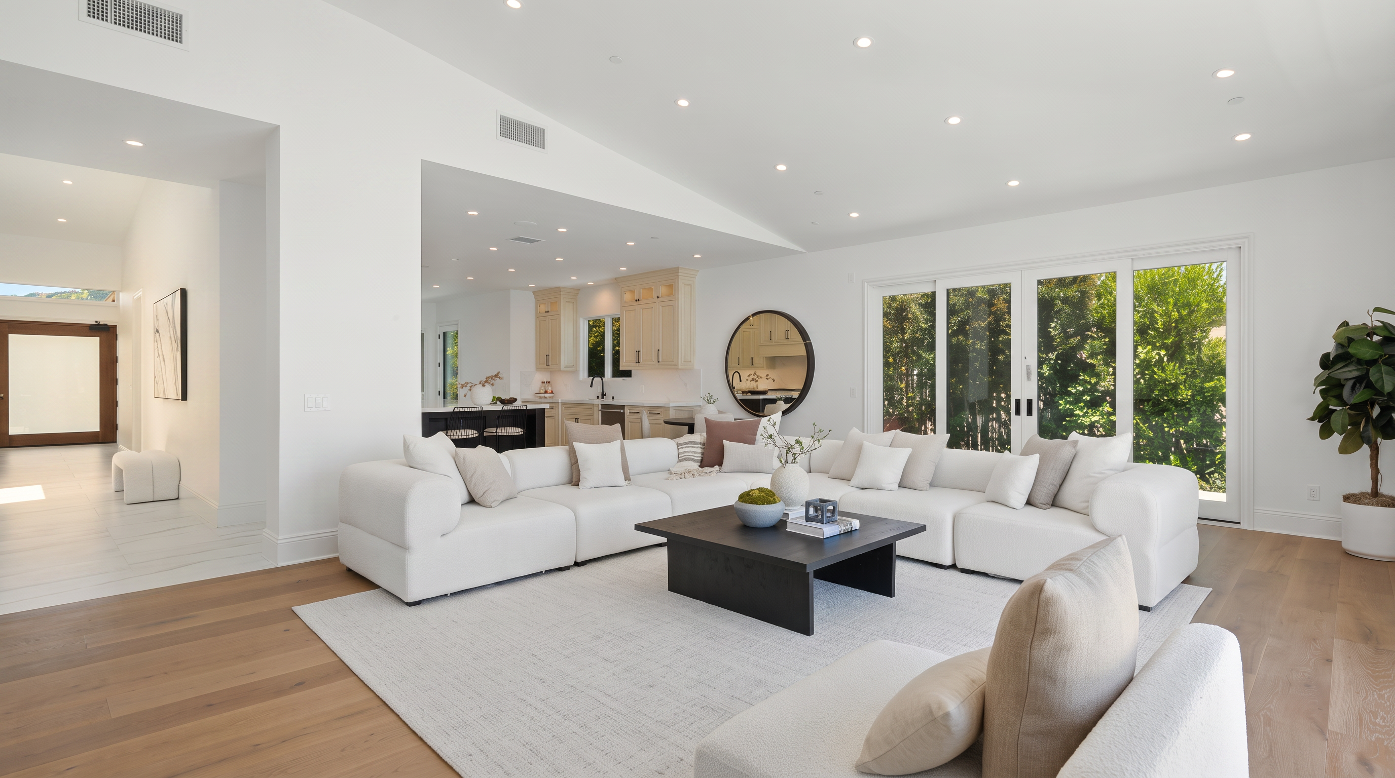 This spacious, open-concept living room features a large, neutral-toned sectional sofa arranged around a dark, low-profile coffee table. The room is bathed in natural light from expansive glass doors that lead to the outdoors, complemented by recessed ceiling lighting. A light-colored area rug anchors the seating area, while a glimpse of the adjacent kitchen and a circular wall mirror add depth and modern elegance to the bright, airy space.