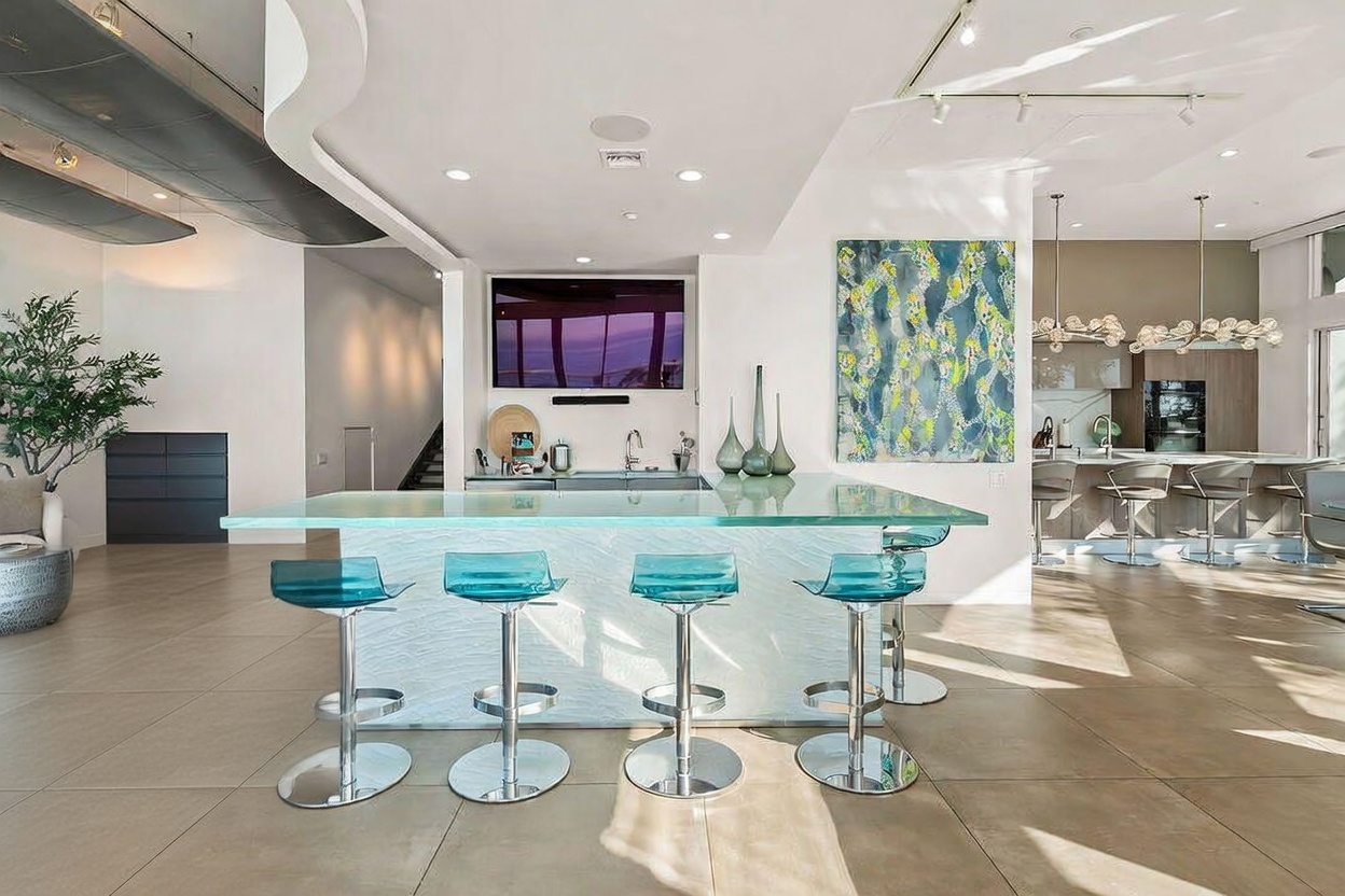 This modern, open-concept kitchen features a prominent central island with a glass countertop and four translucent blue bar stools. The space is bright and airy, showcasing a large abstract painting on the wall and a television mounted above a built-in bar area. In the background, a secondary kitchen island with additional seating and contemporary pendant lighting creates a seamless flow between the entertainment and culinary zones.