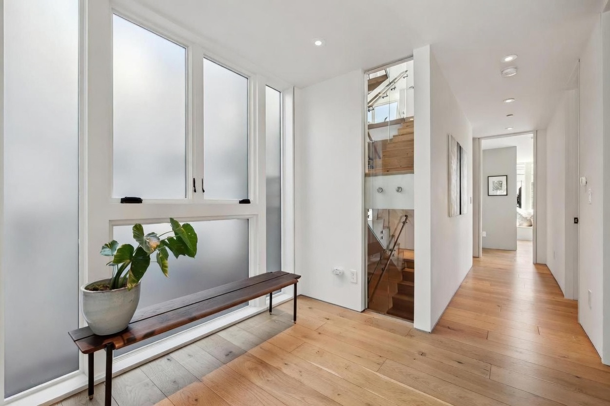 This bright, modern entryway features light wood flooring and crisp white walls, creating an airy and minimalist atmosphere. A long, dark wood bench sits beneath large windows with frosted glass, while a glass-enclosed staircase serves as a striking architectural focal point. The perspective looks down a clean hallway, emphasizing the open, contemporary design of the home.