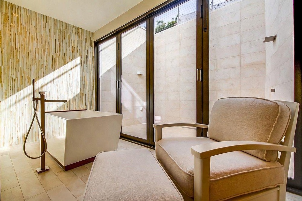 This luxurious bathroom space features a deep, freestanding soaking tub positioned next to a textured, light-toned accent wall. A comfortable armchair with an ottoman sits in the foreground, creating a relaxing lounge area adjacent to large glass doors that open to an outdoor space. The room is bathed in natural light, highlighting the clean lines and neutral color palette of the modern design.