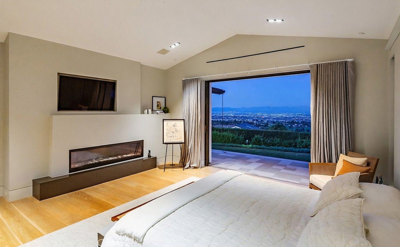 This spacious primary bedroom features a minimalist aesthetic with light wood flooring and a neutral color palette. A wide, linear fireplace is set into a prominent wall feature below a mounted television, while a large opening with sliding glass doors provides a seamless transition to an outdoor patio with expansive city views. The room is furnished with a plush white bed and a comfortable accent chair, creating a serene and luxurious atmosphere.