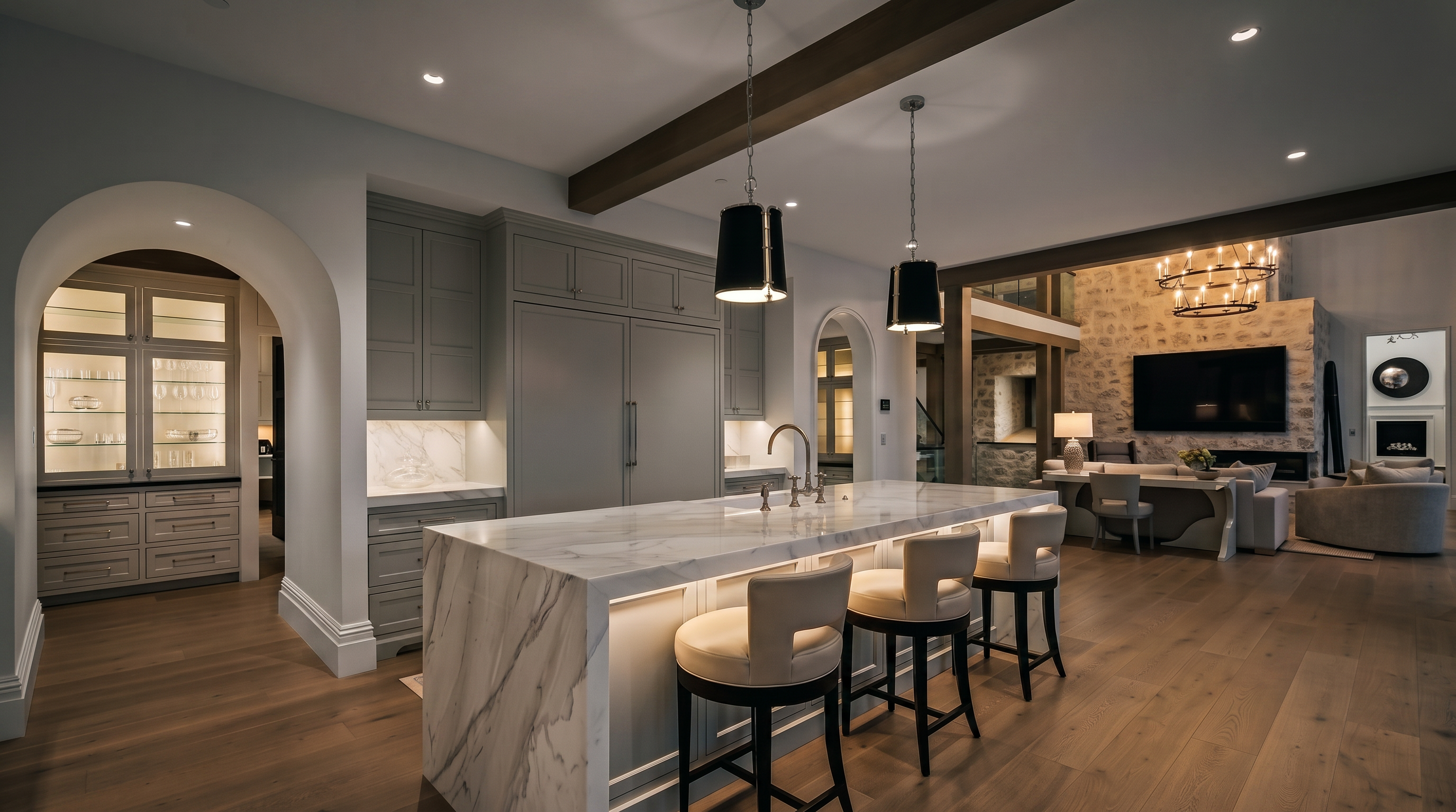 This sophisticated open-concept kitchen features a large central island with a waterfall-edge stone countertop and three upholstered bar stools. The space is defined by light-toned wood flooring, soft gray cabinetry, and a prominent wooden ceiling beam that adds architectural character. In the background, an arched entryway leads to a secondary room, while a living area with a stone-clad chimney breast and a large television completes the expansive, airy layout.