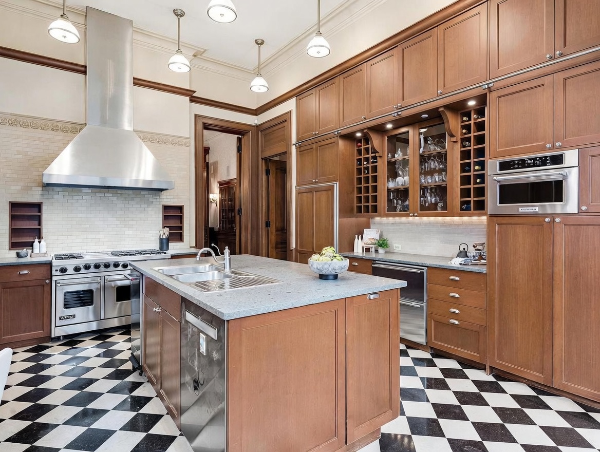 This spacious kitchen features warm-toned cabinetry, a large central island with a stone countertop, and a professional-grade stainless steel range with a prominent vent hood. The room is anchored by a classic black-and-white checkered floor and illuminated by multiple pendant lights hanging from the ceiling. A built-in wine rack and integrated microwave add functional elegance to the traditional design, creating a sophisticated and inviting culinary space.