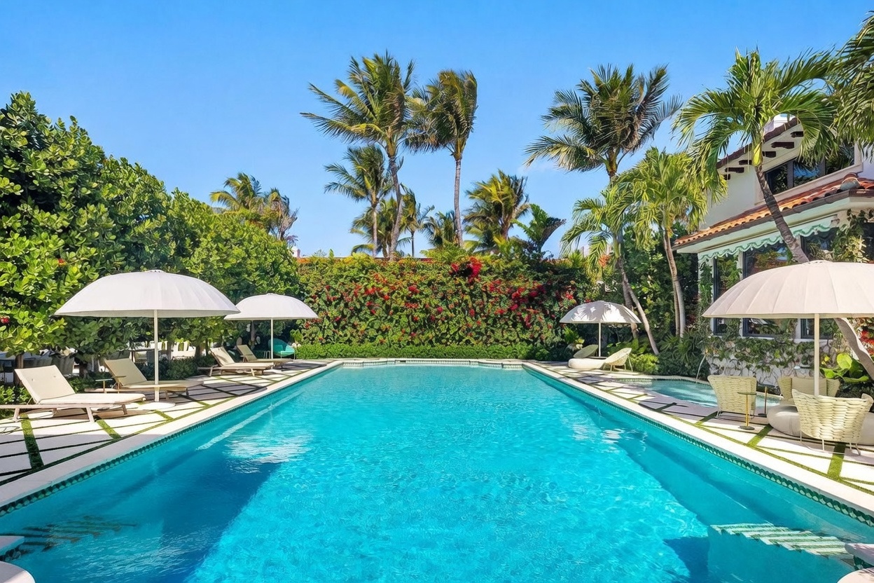 This vibrant outdoor space features a large, rectangular swimming pool as the centerpiece, surrounded by a paved deck with lounge chairs and white umbrellas. A lush, dense hedge covered in blooming red flowers provides a natural privacy screen, while tall palm trees rise against a clear blue sky. The scene captures a serene, resort-like atmosphere perfect for relaxation and outdoor entertaining.