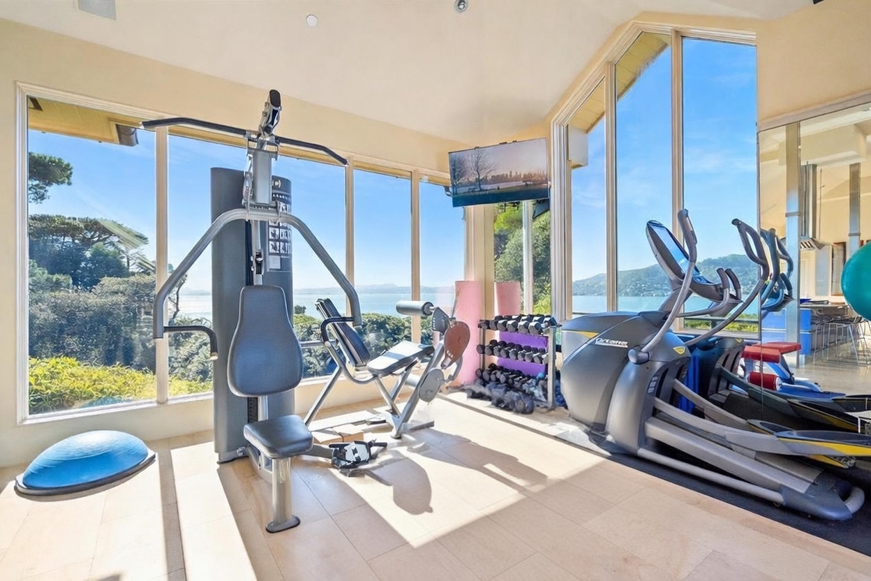 This bright home gym features a comprehensive setup including a multi-station weight machine, an elliptical trainer, a weight bench, and a rack of dumbbells. The room is surrounded by expansive windows that provide panoramic views of the water and surrounding landscape, creating an airy and inspiring workout environment. A blue exercise mat anchors the equipment, while a wall-mounted television provides entertainment during exercise sessions.