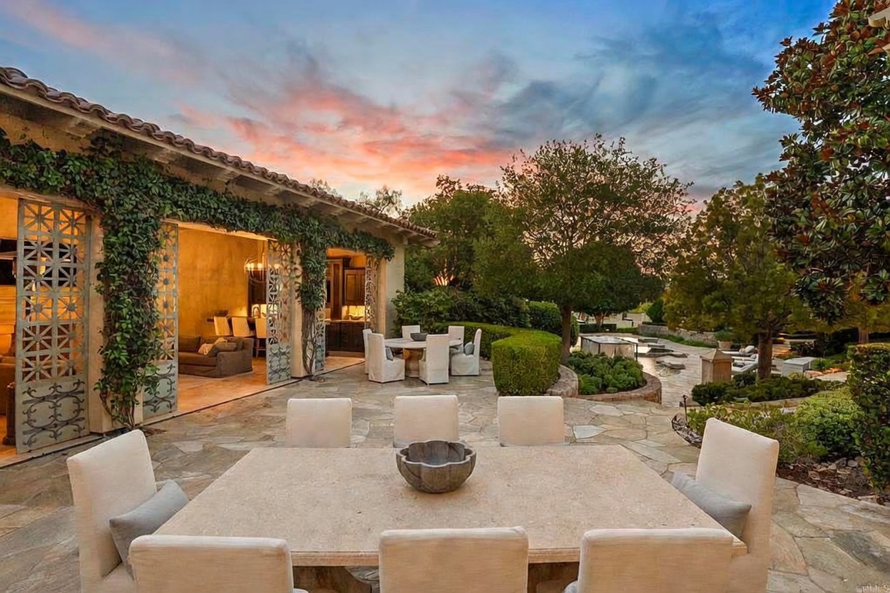 This is a beautifully designed outdoor patio area, perfect for entertaining or relaxing. The space features a large stone dining table with comfortable chairs, set on a stone patio. Lush greenery and mature trees surround the area, creating a private and serene atmosphere, while the open-air structure with climbing vines adds architectural interest and a touch of elegance.