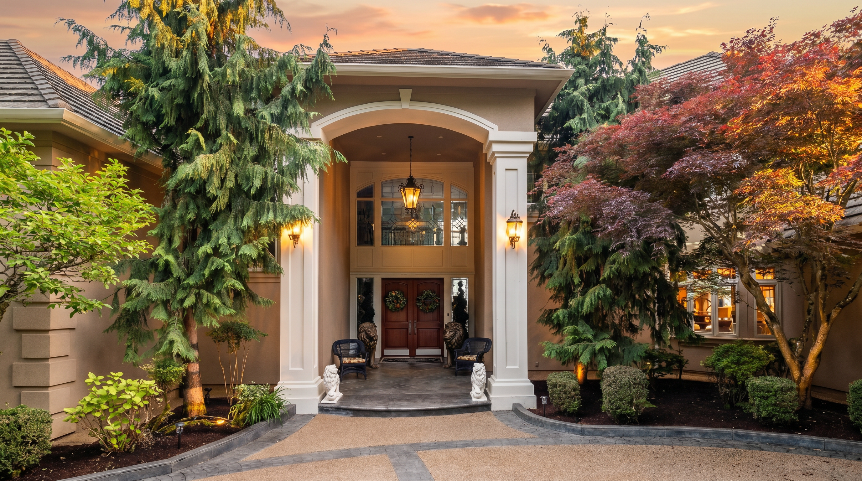 This grand entryway features a covered porch with tall white columns and a decorative arched ceiling. The double front doors are adorned with seasonal wreaths and flanked by two large lion statues, creating an imposing and elegant facade. The paved walkway leads directly to the entrance, framed by lush evergreen trees and manicured landscaping.
