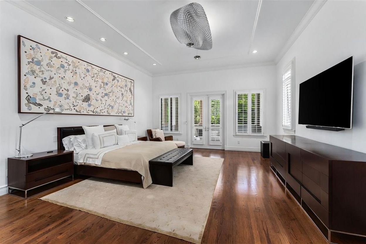 This spacious primary bedroom features rich dark wood flooring and a large, neutral-toned area rug centered under a king-sized bed. A prominent, expansive piece of abstract art hangs above the headboard, while a sleek dark wood dresser sits opposite the bed beneath a wall-mounted television. The room is illuminated by a unique, textured sculptural light fixture and benefits from natural light streaming through French doors flanked by plantation shutters.