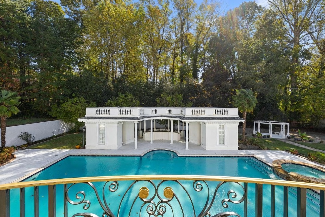 This elevated perspective captures a luxurious backyard oasis featuring a large, rectangular swimming pool with an integrated circular spa. In the background, a white, classical-style pool house with a central open-air bar area and symmetrical wings sits against a backdrop of lush, mature trees. The foreground is framed by an ornate, gold-accented wrought iron railing, providing a sophisticated vantage point overlooking the serene outdoor living space.