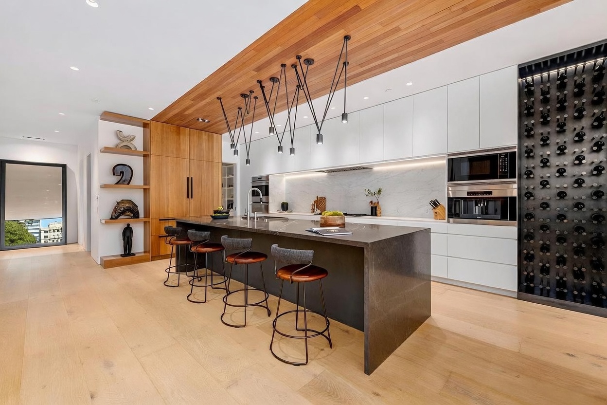 This modern, high-end kitchen features a large central island with dark stone countertops and four bar stools, set against a backdrop of sleek white cabinetry and light wood paneling. A unique, multi-pendant light fixture hangs above the island, while a built-in wine rack occupies the far wall. The space is bright and open, with light wood flooring and a minimalist aesthetic that emphasizes clean lines and premium finishes.