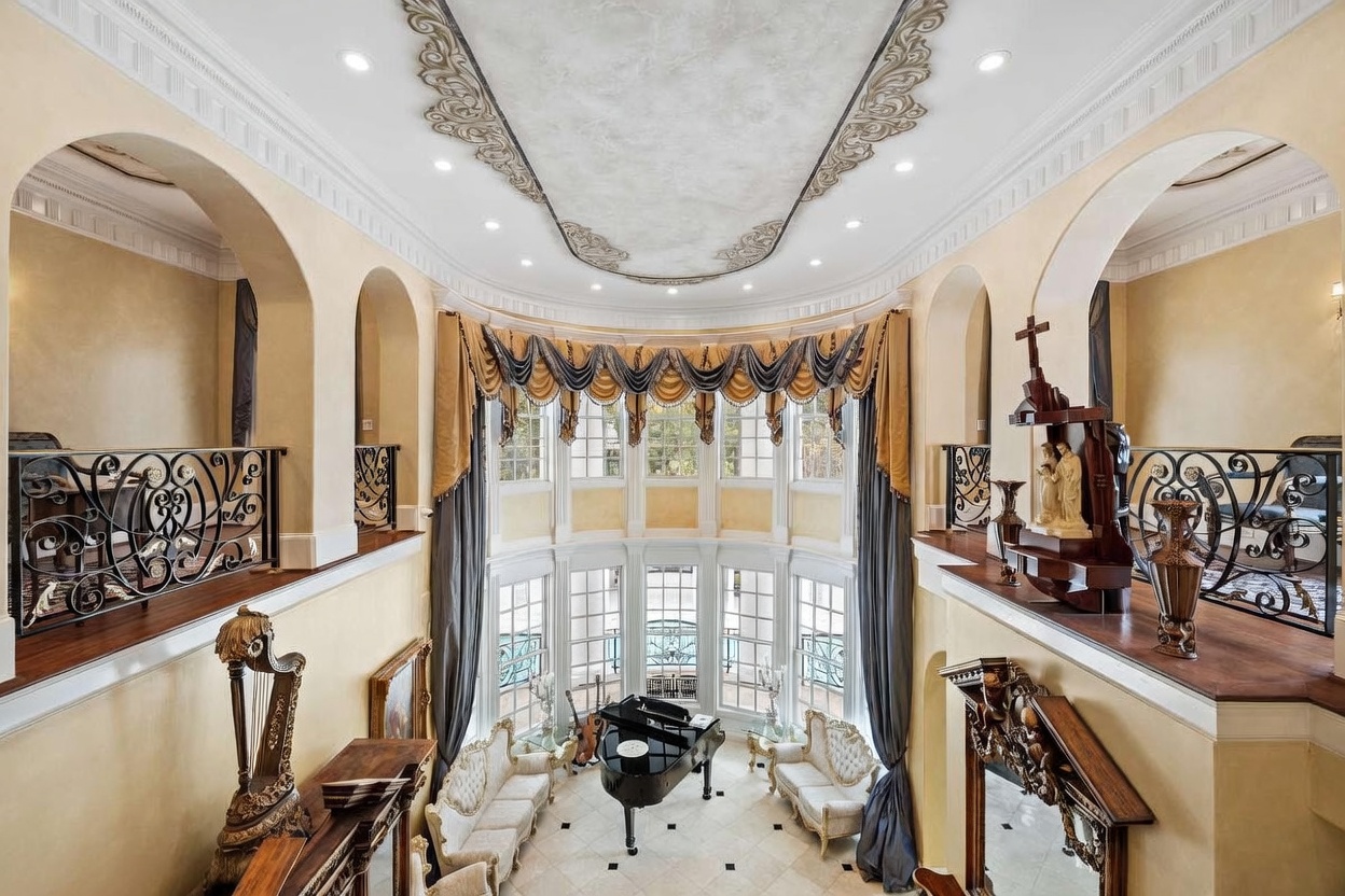 This grand, two-story living room features a dramatic, curved wall of tall windows draped in elegant, multi-layered curtains. The space is anchored by a central black grand piano and ornate, light-colored seating, with a decorative ceiling medallion and intricate iron railings on the upper-level balconies. The perspective is captured from an elevated vantage point, emphasizing the opulent, formal architectural style and the expansive, airy volume of the room.