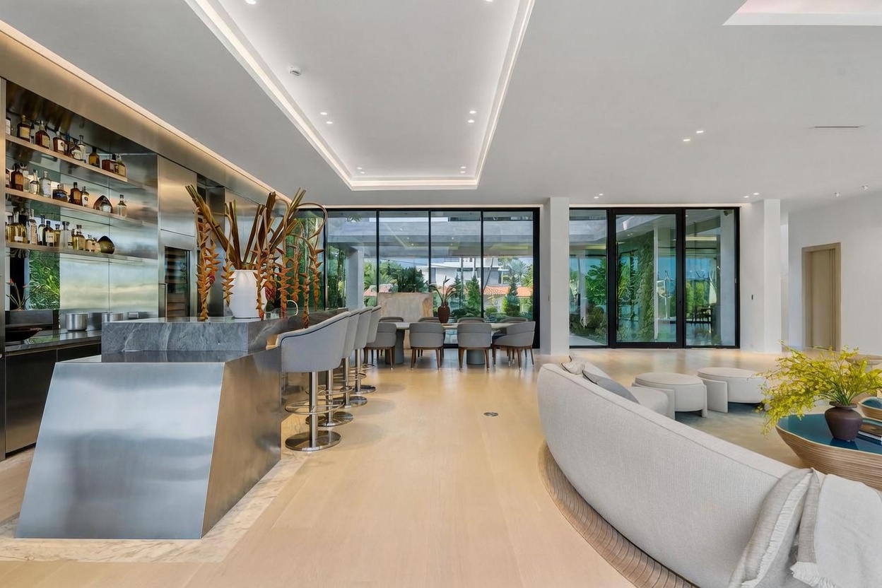 This expansive, open-concept living space features a sleek, modern bar area with a metallic base and stone countertop, complemented by a row of grey bar stools. The room flows seamlessly into a dining area and is framed by large glass walls that provide an unobstructed view of the outdoor landscape. The interior is characterized by light wood flooring, a minimalist aesthetic, and a curved sofa that anchors the seating area, creating a sophisticated and airy atmosphere.