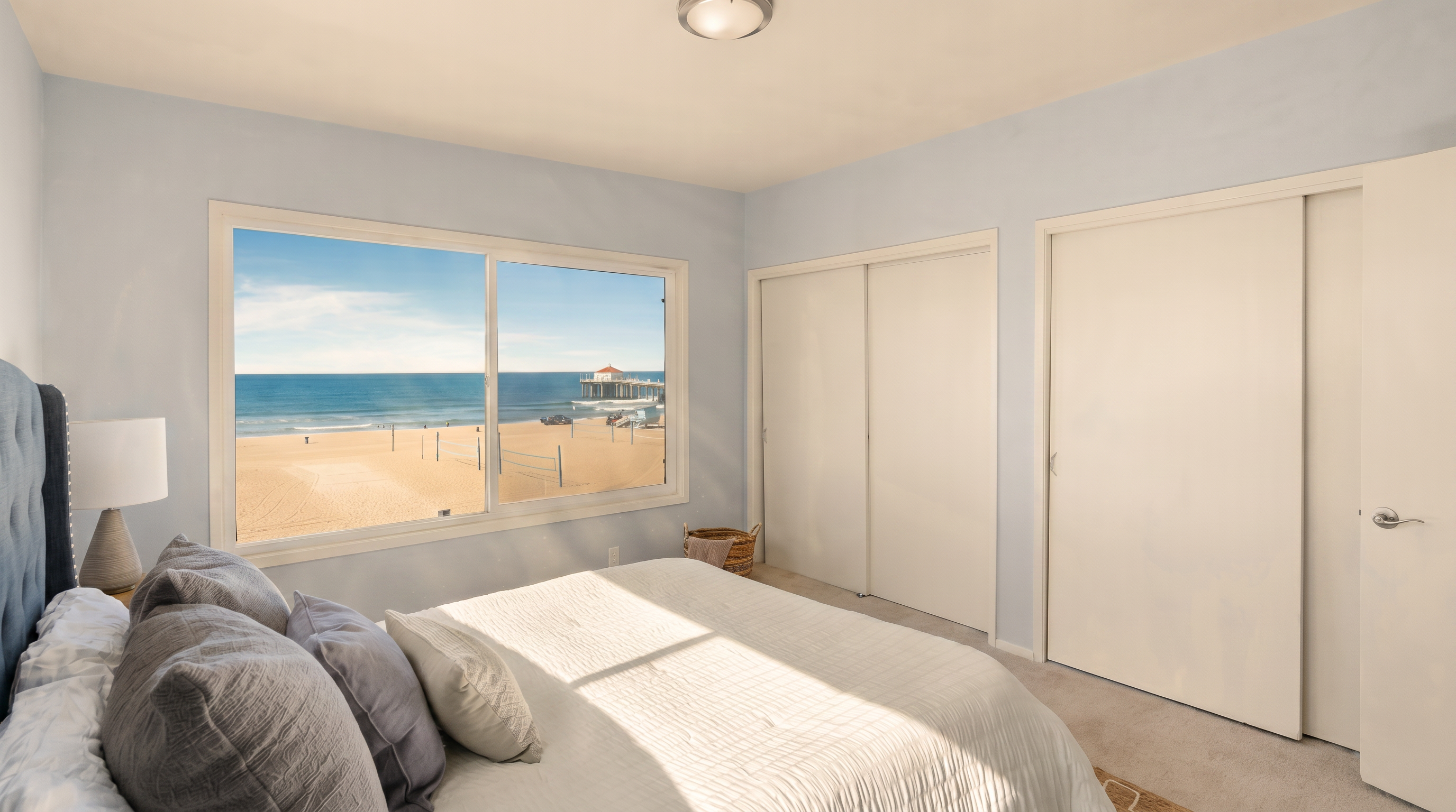This bright bedroom features a neutral color palette and a large window that provides a stunning, direct view of the beach and ocean. The room is furnished with a bed dressed in light-colored linens and a bedside table with a lamp, while white sliding closet doors provide ample storage. The space offers a serene and airy atmosphere, emphasizing the coastal location.