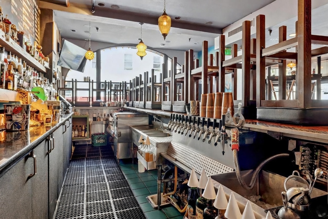 This image captures the interior of a commercial bar area, featuring a long service counter equipped with multiple beer taps and a stainless steel sink station. The space is organized with chairs placed upside down on the bar top, suggesting the establishment is closed or in a cleaning phase. Decorative pendant lights hang from the ceiling, and a large window at the back provides natural light, creating a professional and functional atmosphere.