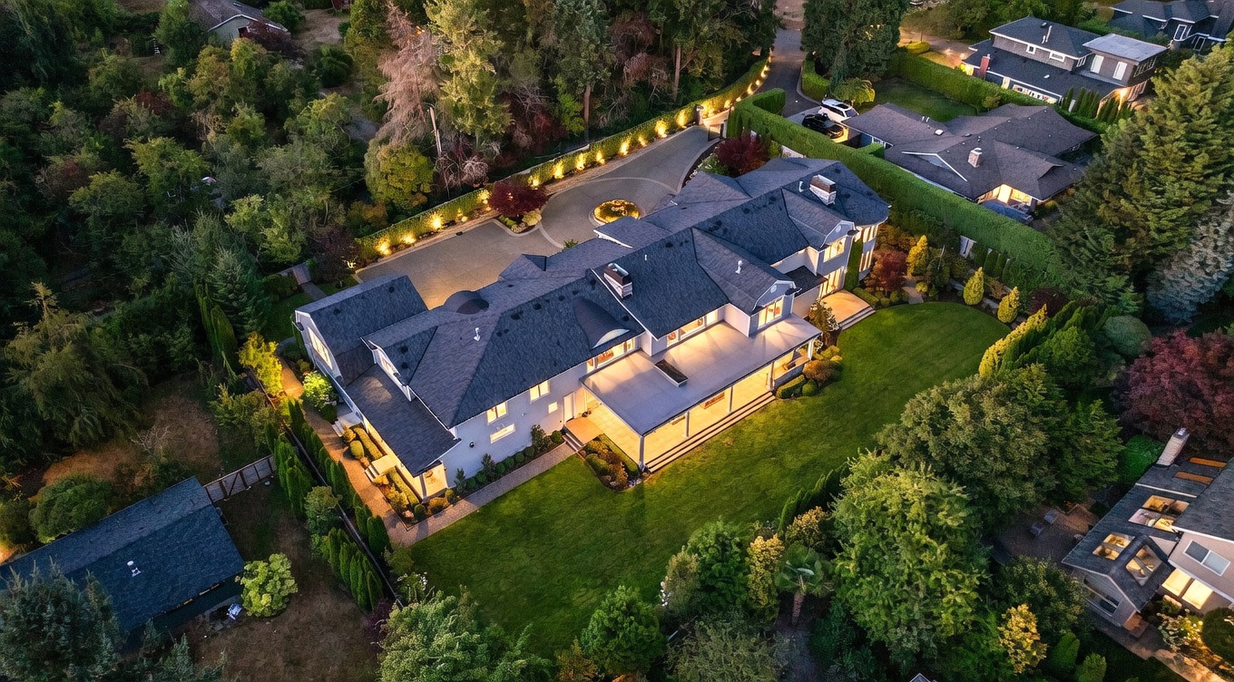 This high-angle aerial view captures a sprawling, multi-story luxury residence at dusk, illuminated by warm exterior lighting. The home features a complex roofline, a large covered patio overlooking a manicured lawn, and a circular driveway with a central fountain feature. The property is surrounded by dense, mature landscaping and tall hedges, providing a sense of privacy and exclusivity.