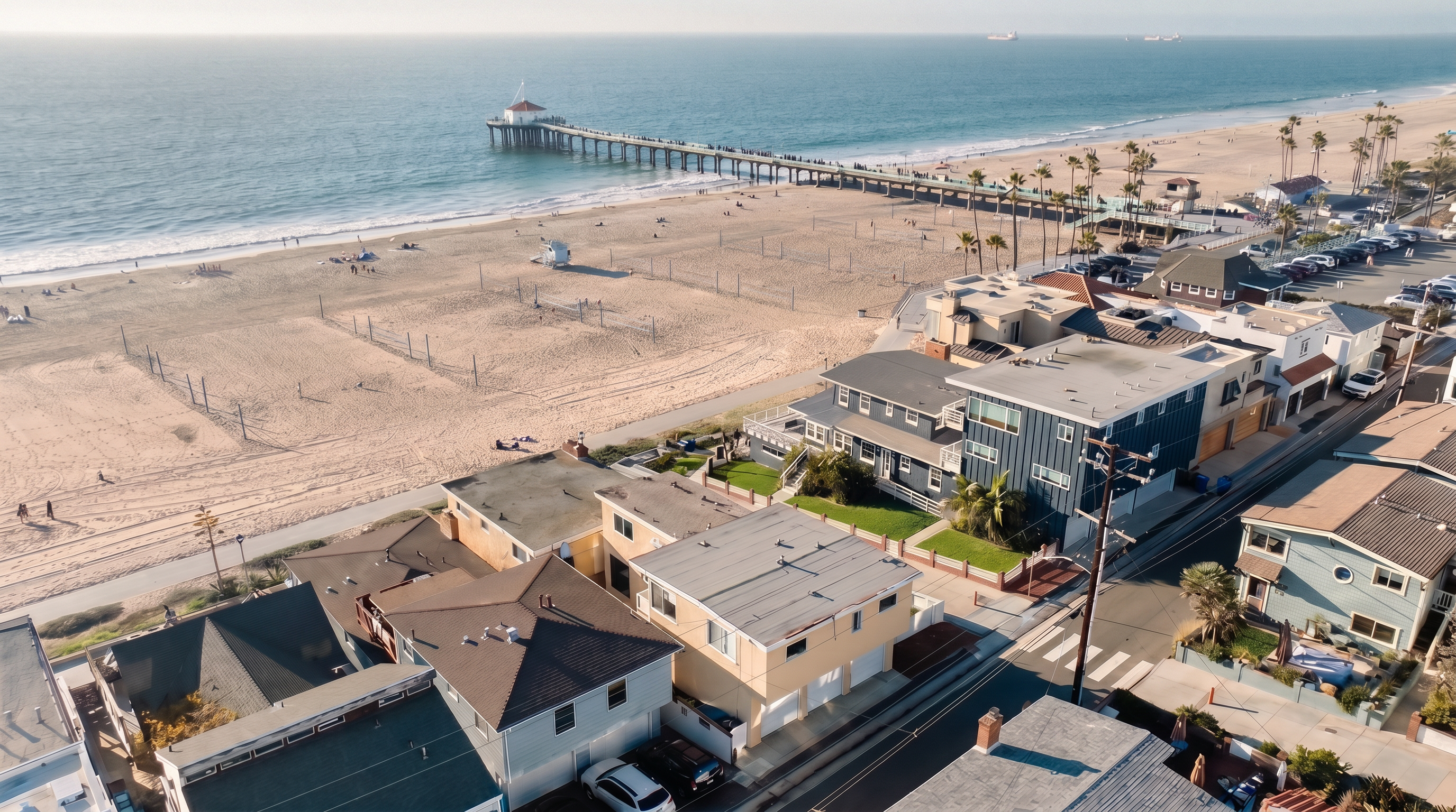This high-angle aerial view captures a prime coastal property situated directly adjacent to a sandy beach and a prominent pier. The multi-story residence features a flat roof and a light-colored exterior, positioned within a dense neighborhood of similar beach-front homes. The perspective emphasizes the property's exceptional proximity to the ocean, highlighting the expansive shoreline and the surrounding community layout.