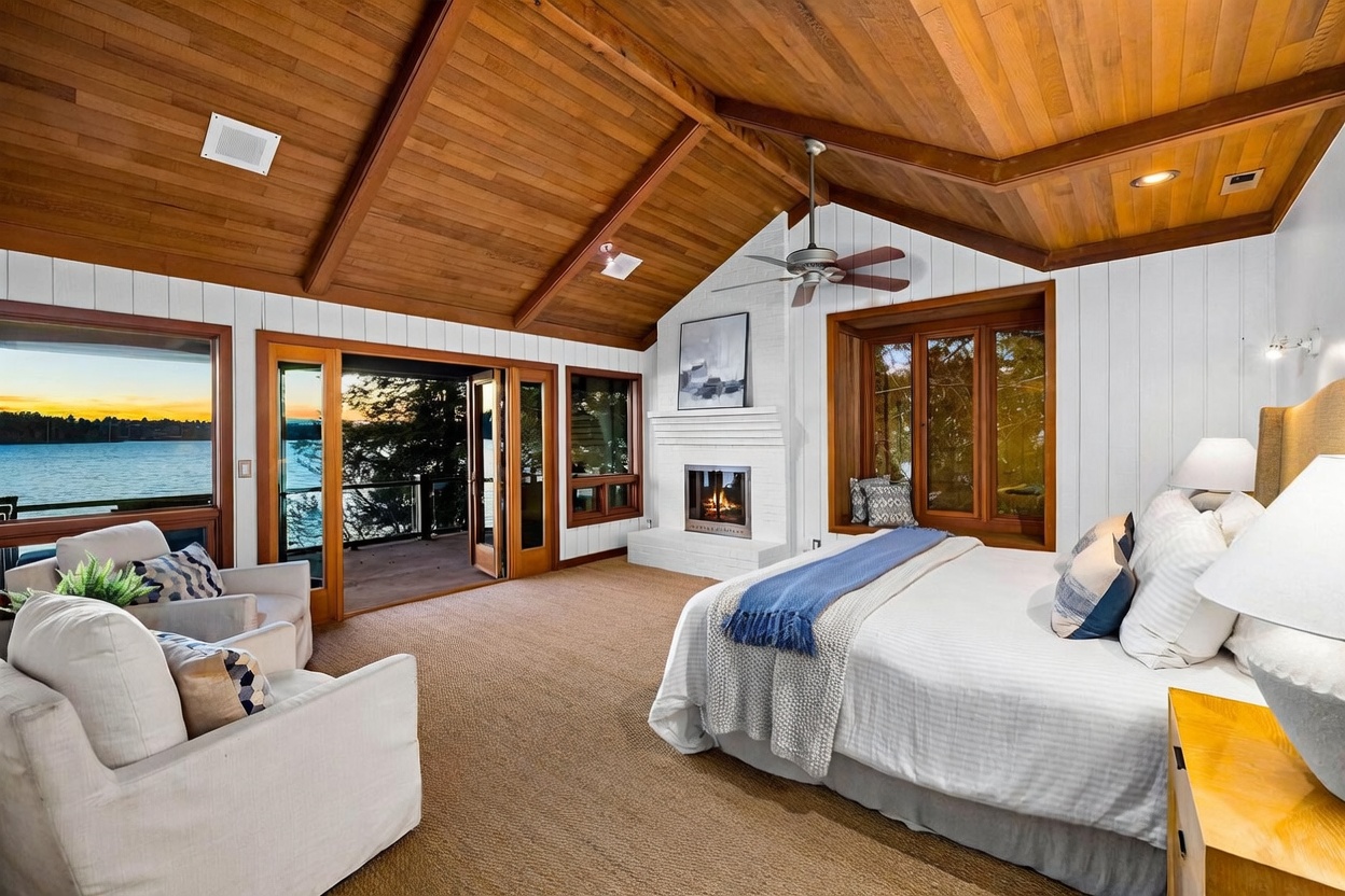 This spacious primary bedroom features a vaulted wood-paneled ceiling, a cozy fireplace, and expansive windows that offer a stunning view of the lake. The room is furnished with a plush bed and a comfortable seating area, creating a serene and luxurious retreat. The combination of natural wood textures, white shiplap walls, and direct access to an outdoor deck highlights the home's elegant, nature-inspired design.
