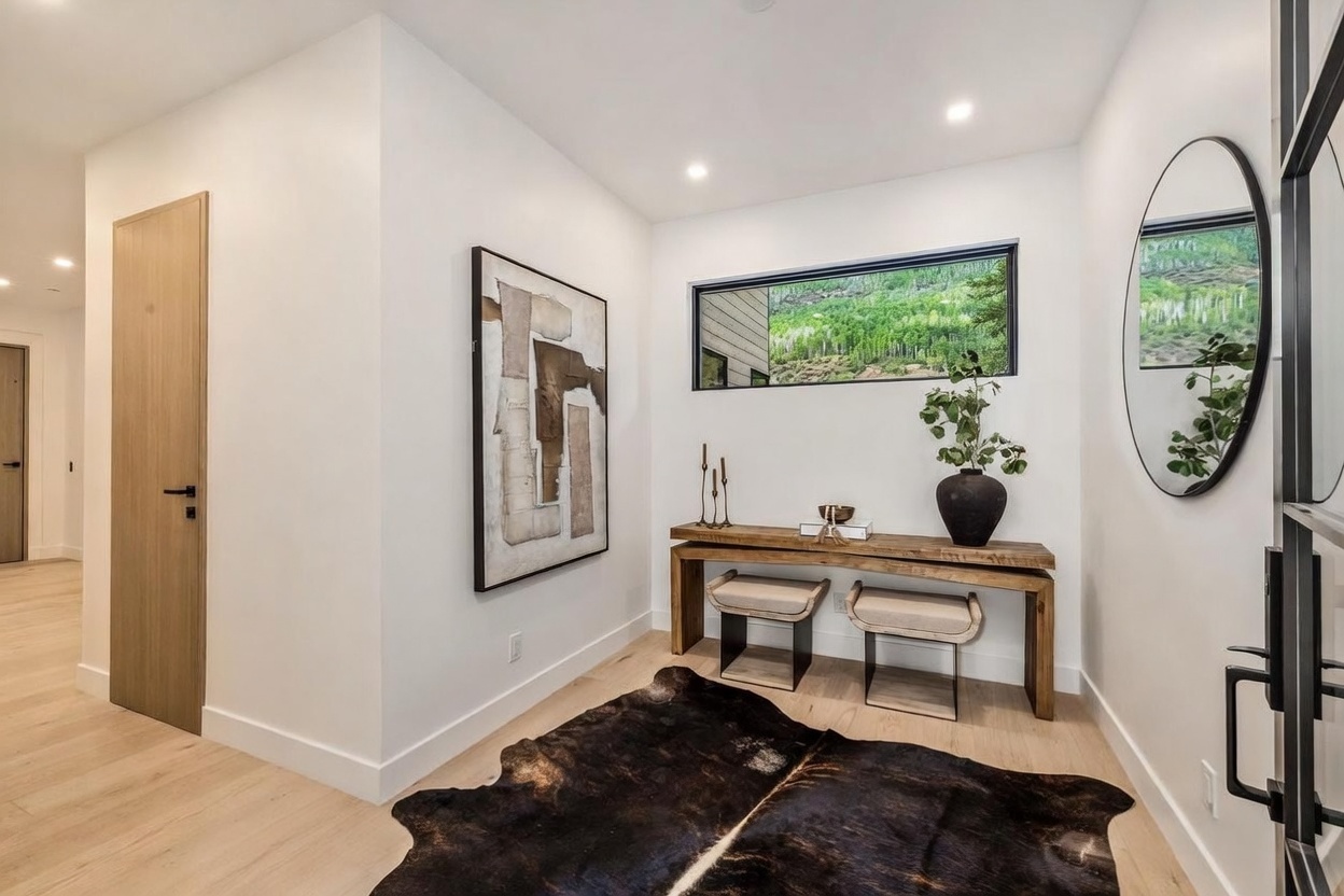 This elegant entryway features light wood flooring and a minimalist aesthetic, anchored by a rustic wooden console table and two upholstered stools. A large, abstract piece of wall art adds a sophisticated focal point, while a circular mirror reflects the natural light streaming through a horizontal window overlooking a lush, wooded landscape. The space is finished with a dark, textured animal hide rug, creating a warm and inviting transition area.