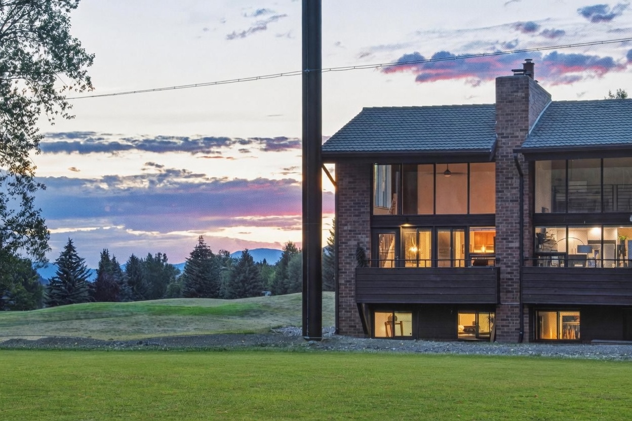 This rear view of a multi-story brick residence showcases a contemporary design with expansive glass walls that offer panoramic views of the surrounding landscape. The home features a prominent dark metal chimney, a private balcony, and a walk-out basement level that opens directly onto a manicured lawn. Set against a dramatic sunset sky with mountain silhouettes in the distance, the property exudes a serene and sophisticated atmosphere.
