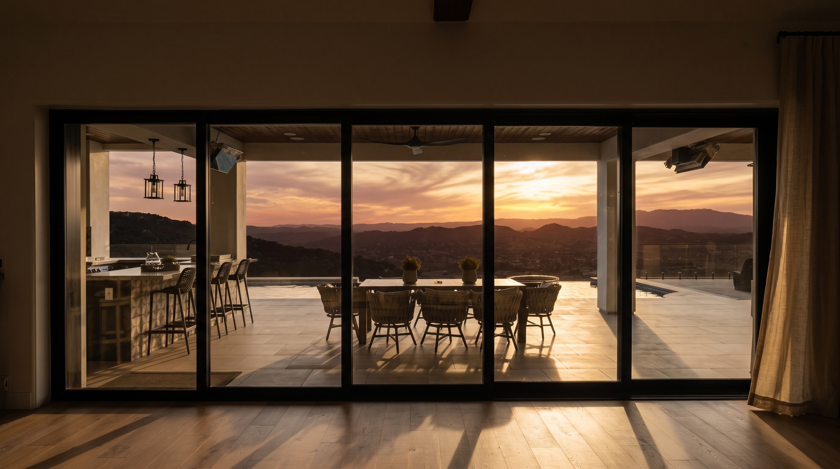 This interior view looks out through expansive floor-to-ceiling black-framed sliding glass doors onto a luxurious outdoor patio area. The outdoor space features a well-equipped kitchen island with bar seating and a dining area, all set against a stunning panoramic backdrop of rolling hills and valley views. The interior itself features light-toned hardwood flooring and a clean, minimalist design, emphasizing a seamless indoor-outdoor living experience.