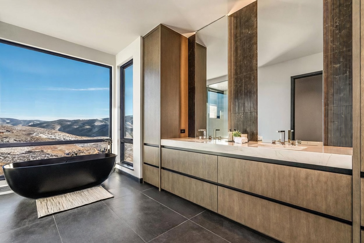 This is a luxurious primary bathroom featuring a modern freestanding black bathtub positioned in front of a large window with mountain views. The bathroom includes a double vanity with sleek wood cabinetry, a marble countertop, and contemporary fixtures. The overall design is minimalist and elegant, emphasizing natural light and high-end finishes.