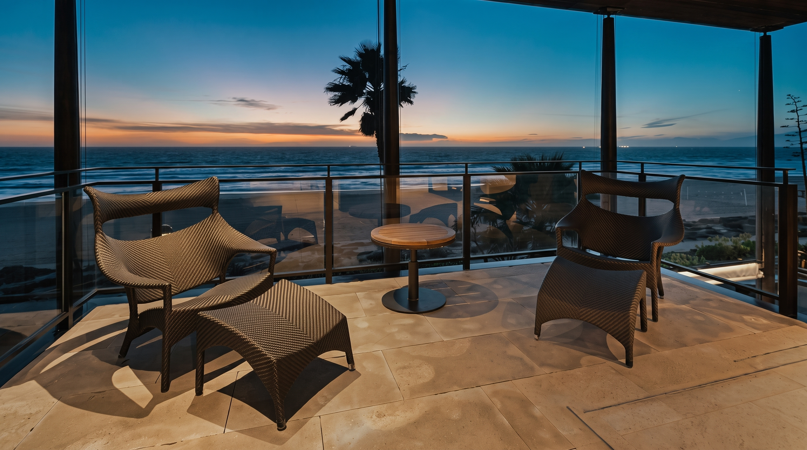 This elevated balcony offers a serene, unobstructed view of the ocean at twilight. The space is furnished with two modern woven chairs, each paired with a matching ottoman and a small central table, set against a backdrop of a glass railing. The scene captures a peaceful coastal atmosphere with the horizon glowing in soft sunset hues.