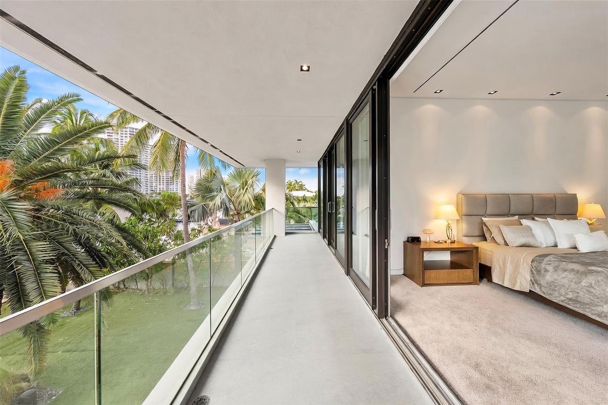 This image captures a spacious, covered balcony that seamlessly connects to a primary bedroom through large sliding glass doors. The balcony features a sleek glass railing, providing an unobstructed view of the lush, tropical greenery and distant city skyline. The bedroom interior is visible, showcasing a neutral-toned bed with plush bedding and warm bedside lighting, creating a serene and luxurious atmosphere.