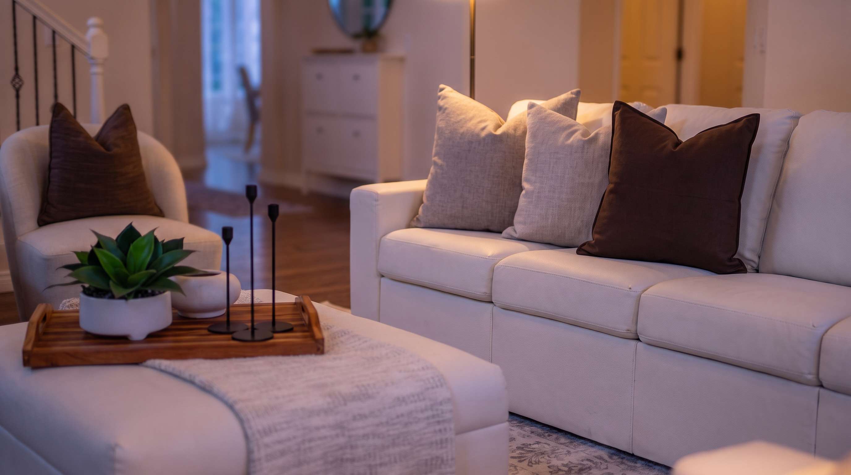 This living room provides an inviting and sophisticated atmosphere, anchored by a large, comfortable white sofa and matching armchair set. The space features elegant design touches including a textured area rug, a central ottoman with a decorative tray, and a warm, ambient glow from the layered lighting fixtures. High ceilings and a prominent staircase contribute to an open and spacious floor plan, making it an ideal setting for gathering and relaxation.