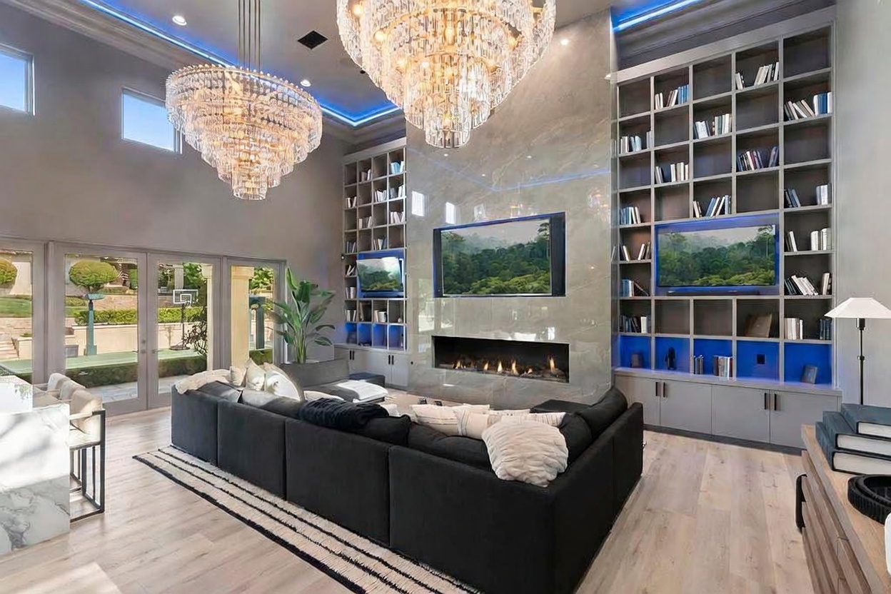 This is a luxurious living room featuring two large crystal chandeliers and a modern fireplace set within a marble wall. The room is flanked by built-in bookshelves with blue accents and multiple televisions. A large sectional sofa anchors the space, complemented by a striped rug and light wood flooring, creating an elegant and inviting atmosphere.
