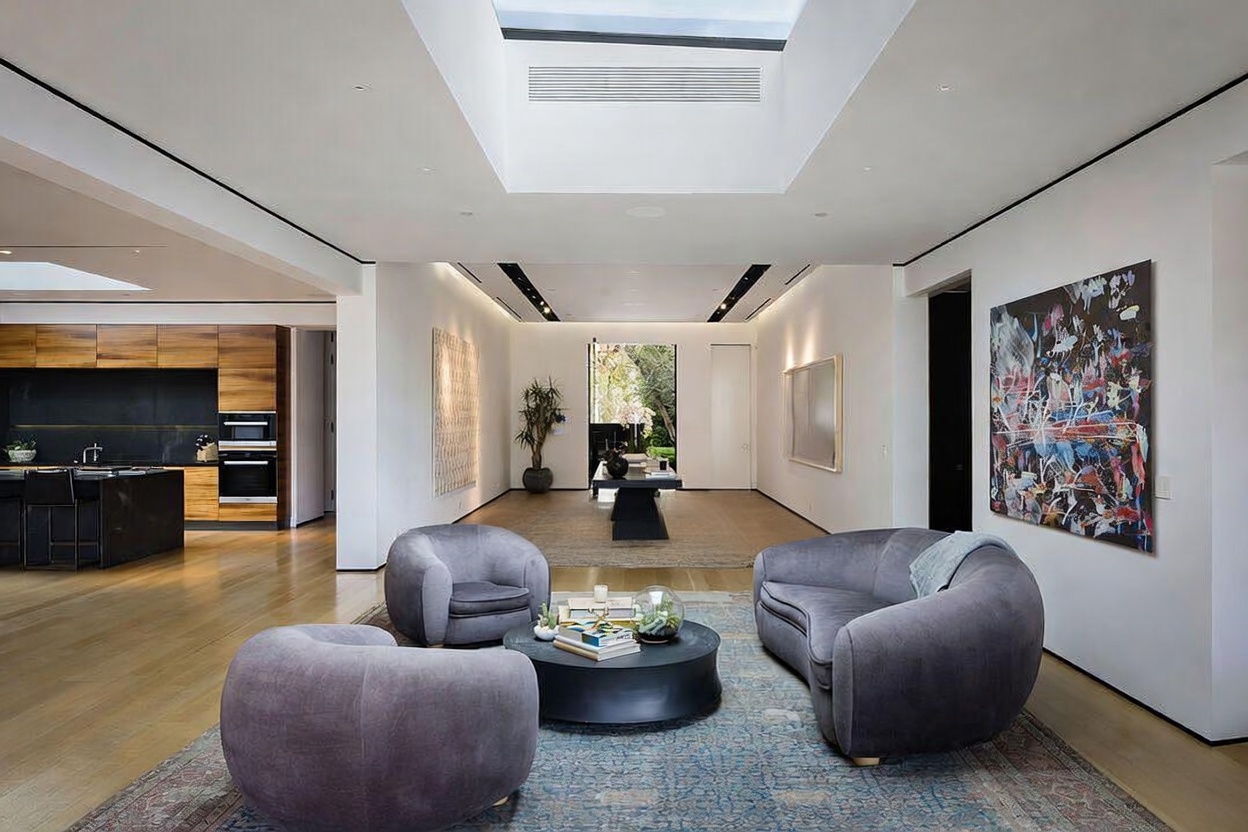 This expansive, open-concept living area features a modern aesthetic with light wood flooring and a dramatic skylight that floods the space with natural light. The room is furnished with a curved sofa and two matching armchairs arranged around a circular coffee table, all set atop a large patterned area rug. To the left, a sleek kitchen with dark cabinetry and wood-toned accents is visible, while a bold, abstract art piece hangs on the right wall, creating a sophisticated and airy atmosphere.