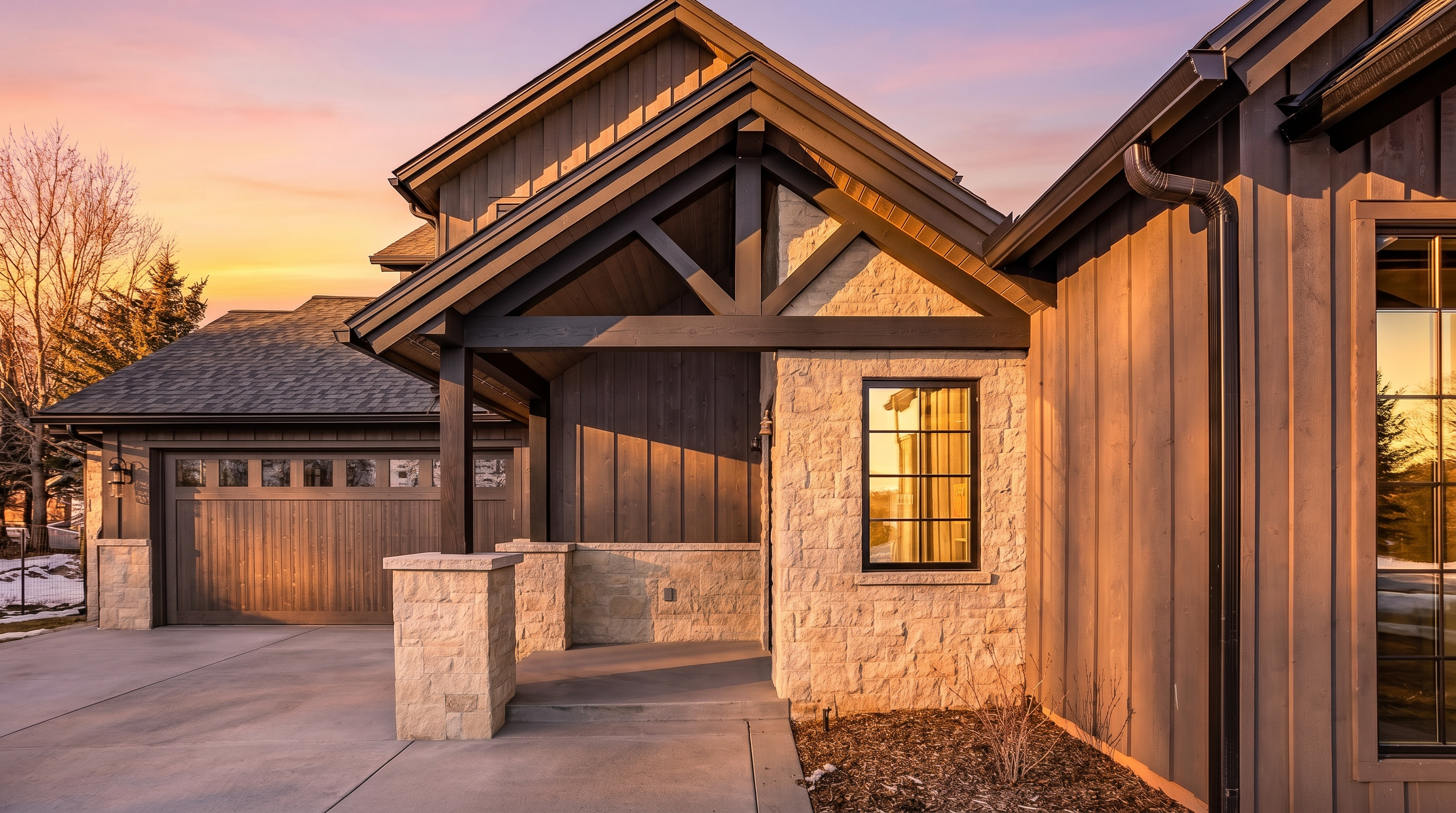 This striking entryway features a blend of rustic stone masonry and dark vertical wood siding, creating a sophisticated modern farmhouse aesthetic. A covered porch with a timber-framed gable roof provides a welcoming focal point, complemented by a multi-pane window and a concrete driveway leading to an attached garage. The clear blue sky and crisp lighting emphasize the high-quality craftsmanship and architectural depth of the home's facade.