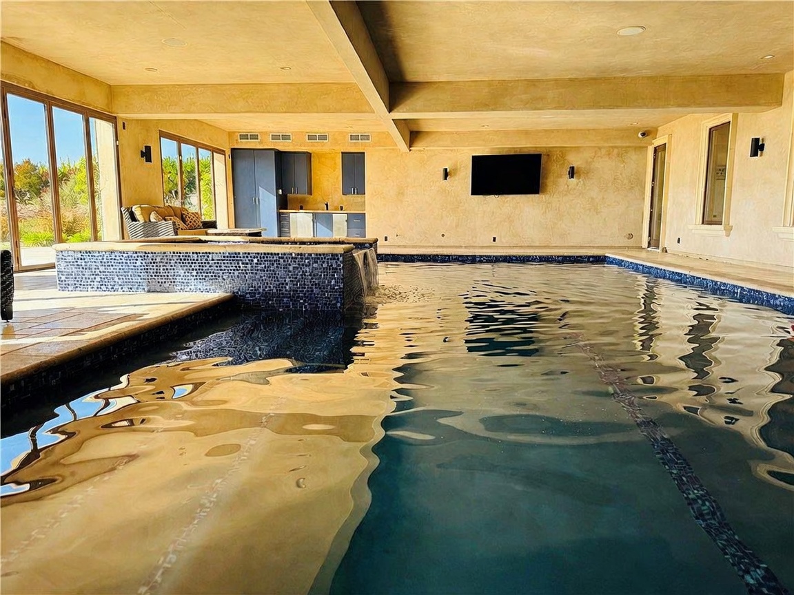 This indoor-outdoor pool area features a large, blue-tiled swimming pool with an integrated spa and a cascading water feature. The space is covered by a high, neutral-toned ceiling and includes a built-in wet bar area with cabinetry in the background, making it an ideal space for entertaining. Large glass doors open to the exterior, allowing natural light to flood the room and providing a seamless transition between the indoor pool and the surrounding landscape.