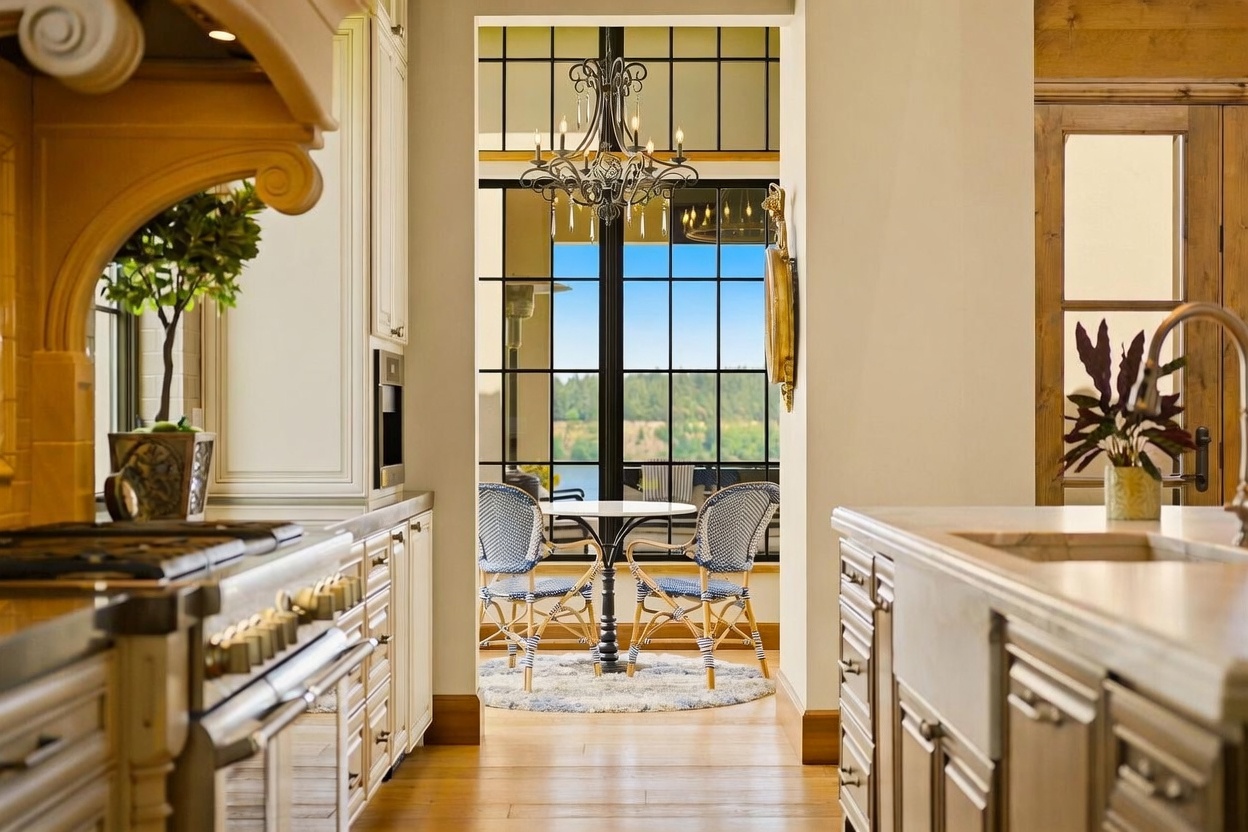 This view from a kitchen looks through a doorway into a bright breakfast nook featuring a round table and patterned chairs. The kitchen area includes light-colored cabinetry, a stove, and a stone countertop, while the transition space is defined by a decorative chandelier and large windows that frame an outdoor view. The overall aesthetic is elegant and inviting, emphasizing a seamless connection between the indoor cooking space and the scenic exterior.