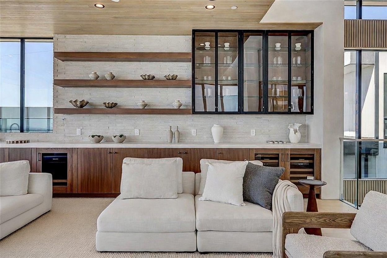 This contemporary living space features a large, light-colored sectional sofa positioned in front of a built-in bar area with wood cabinetry and open shelving. The wall behind the bar is finished with textured stone, complemented by a glass-fronted display cabinet and a wood-paneled ceiling. The perspective is a straight-on, eye-level shot that highlights the clean lines and sophisticated, neutral aesthetic of the room.