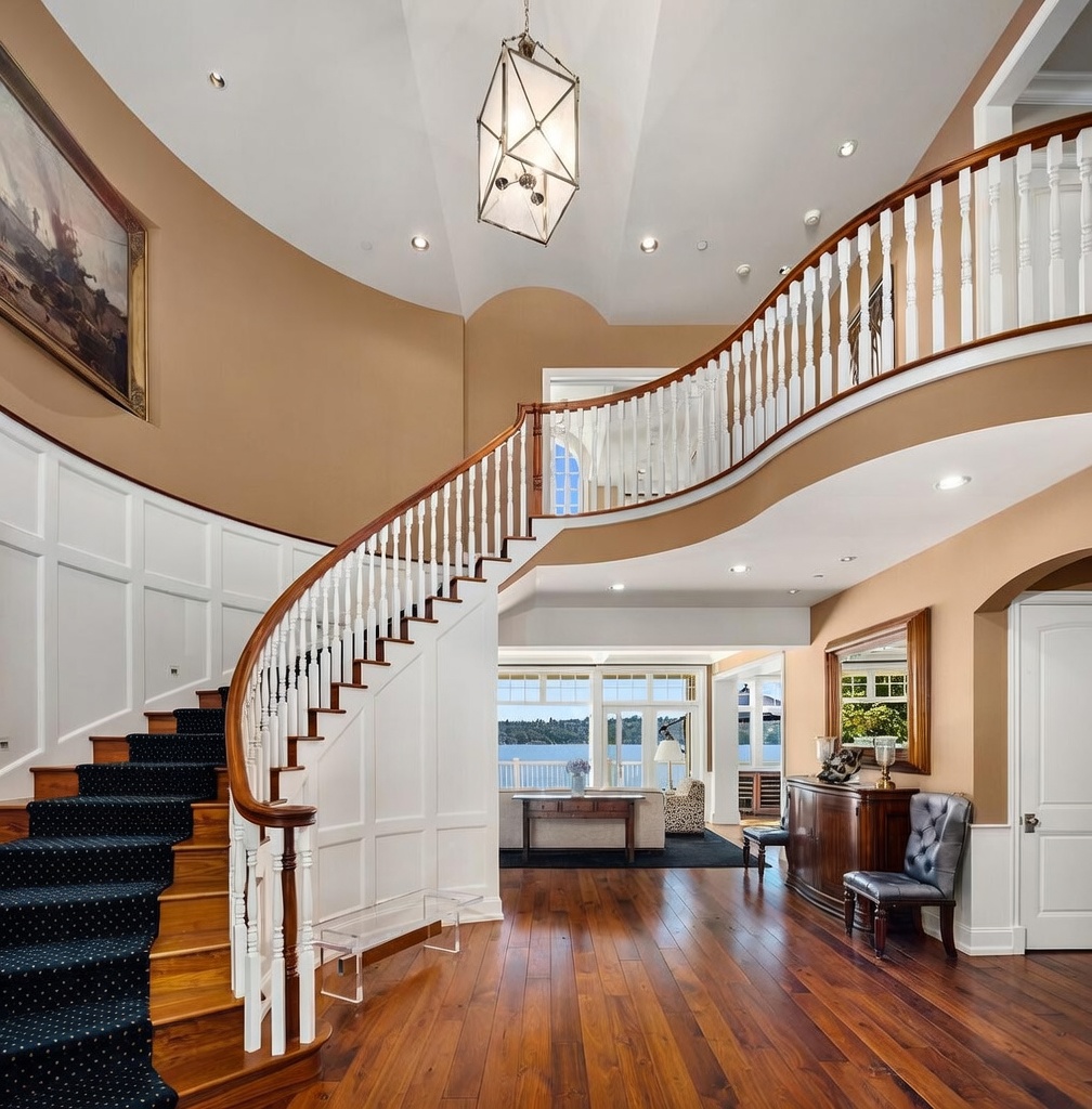 This grand entryway features a sweeping, curved staircase with a dark blue patterned runner and white balusters, serving as the focal point of the home. The space is characterized by high ceilings, warm hardwood flooring, and a sophisticated lantern-style chandelier that adds a touch of elegance. Through the open archway, a glimpse of a bright living area with a scenic water view creates an inviting and luxurious atmosphere.