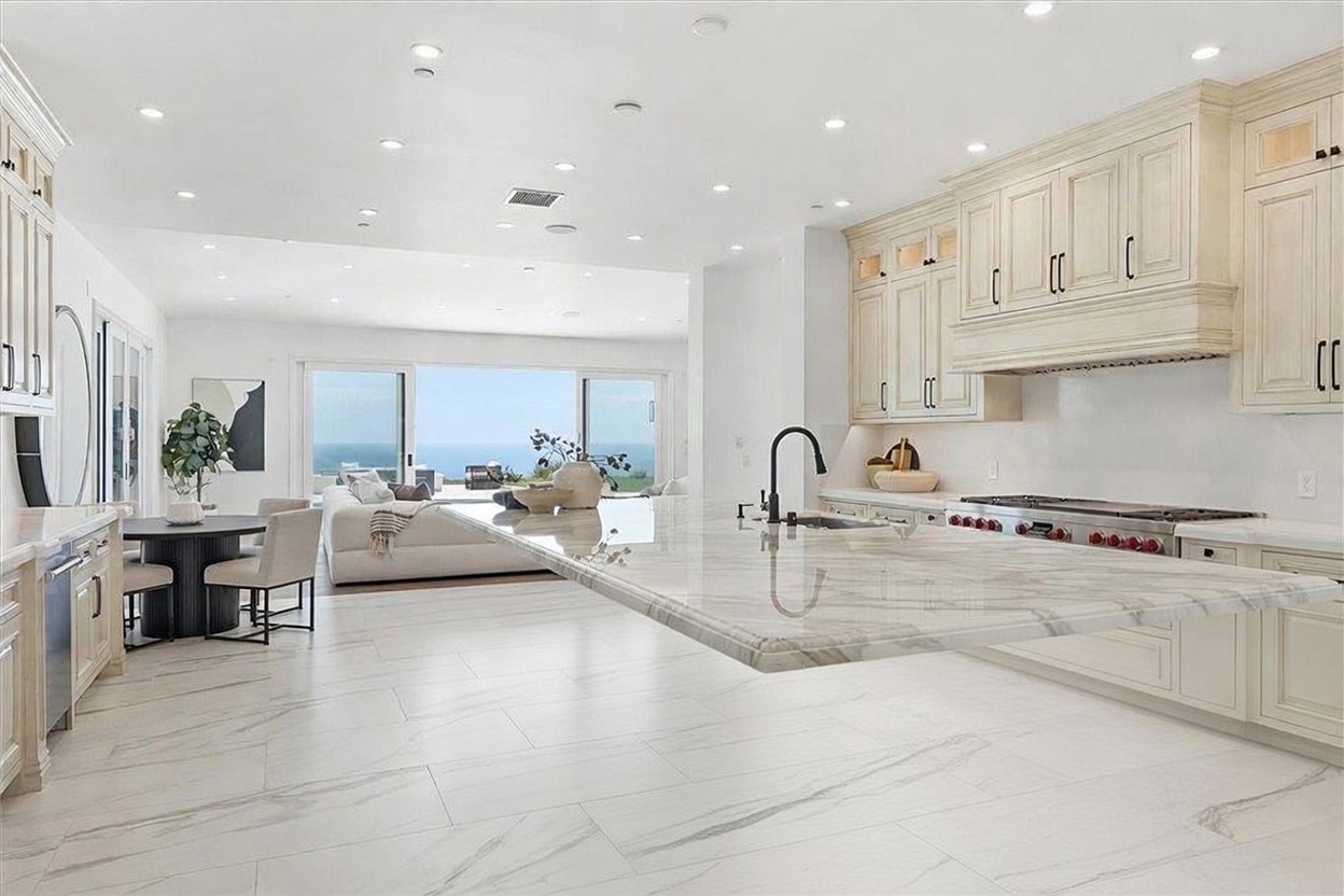 This bright, open-concept kitchen features a large central island with a dark base and a light-toned stone countertop, complemented by light-colored cabinetry and a professional-grade range. The space flows seamlessly into a dining area and living room, which are framed by large glass sliding doors that reveal a panoramic ocean view. The overall aesthetic is sophisticated and airy, emphasizing a clean, modern design with high-end finishes.