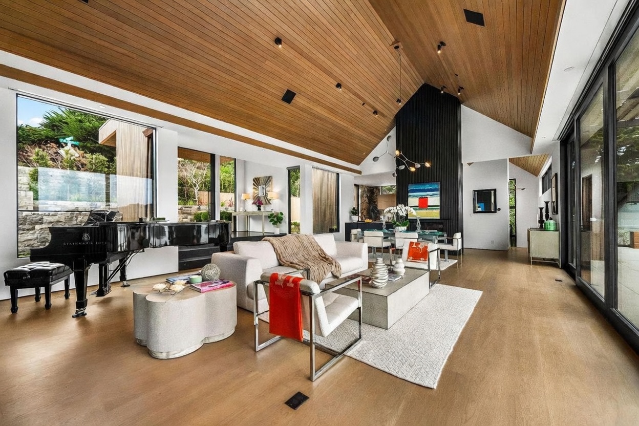 This expansive, open-concept living area features a dramatic vaulted wood-paneled ceiling and a striking black chimney breast that serves as a focal point. The space is furnished with a contemporary sofa, a pair of modern armchairs, and a grand piano, all set against a backdrop of large windows that invite natural light and outdoor views. The light wood flooring and neutral color palette create a sophisticated, airy atmosphere perfect for entertaining.