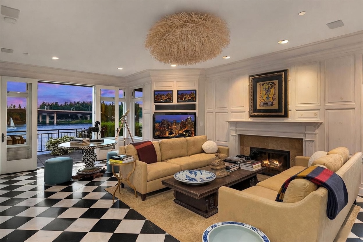 This elegant living room features a striking black-and-white checkered floor and a unique, textured spherical chandelier. The space is anchored by a fireplace with a traditional mantel and a framed piece of art above, flanked by built-in cabinetry and a television. Large glass doors open onto a balcony, providing a scenic view of the water and a bridge at dusk.