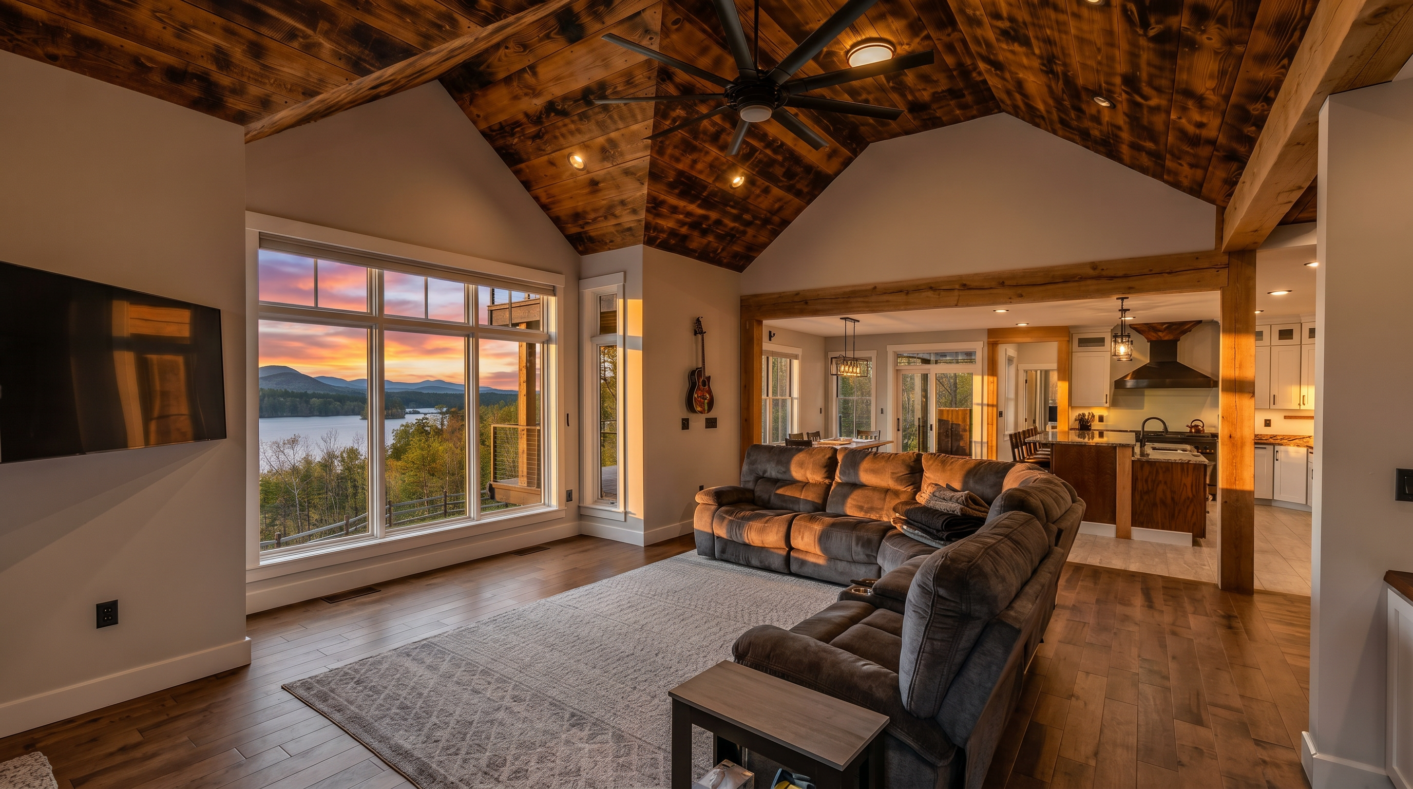 This spacious living room features a vaulted ceiling with exposed wood paneling and a large ceiling fan, creating a rustic yet refined atmosphere. A large sectional sofa sits atop a textured area rug, facing a wall-mounted television and expansive windows that provide a panoramic view of the lake and surrounding mountains. The open-concept design flows seamlessly into the adjacent kitchen and dining area, emphasizing a bright and airy layout.