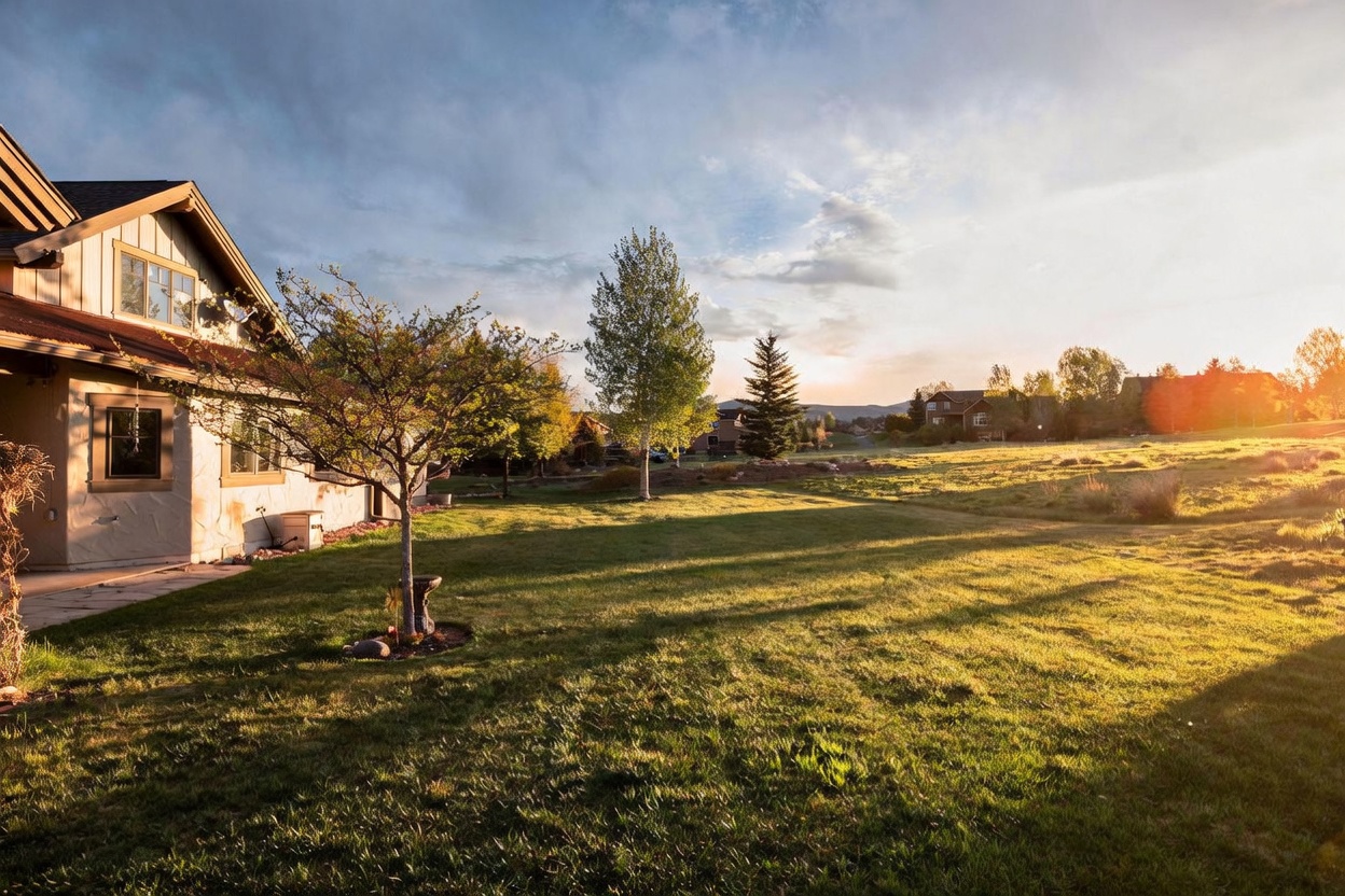 This image captures the rear exterior of a home during the golden hour, showcasing a spacious, well-manicured lawn that stretches toward a scenic mountain backdrop. The house features a light-colored stucco facade with a prominent roofline and large windows, while a small tree and birdbath add charm to the immediate yard. The warm, low-angle sunlight casts long, dramatic shadows across the grass, creating an inviting and peaceful atmosphere.