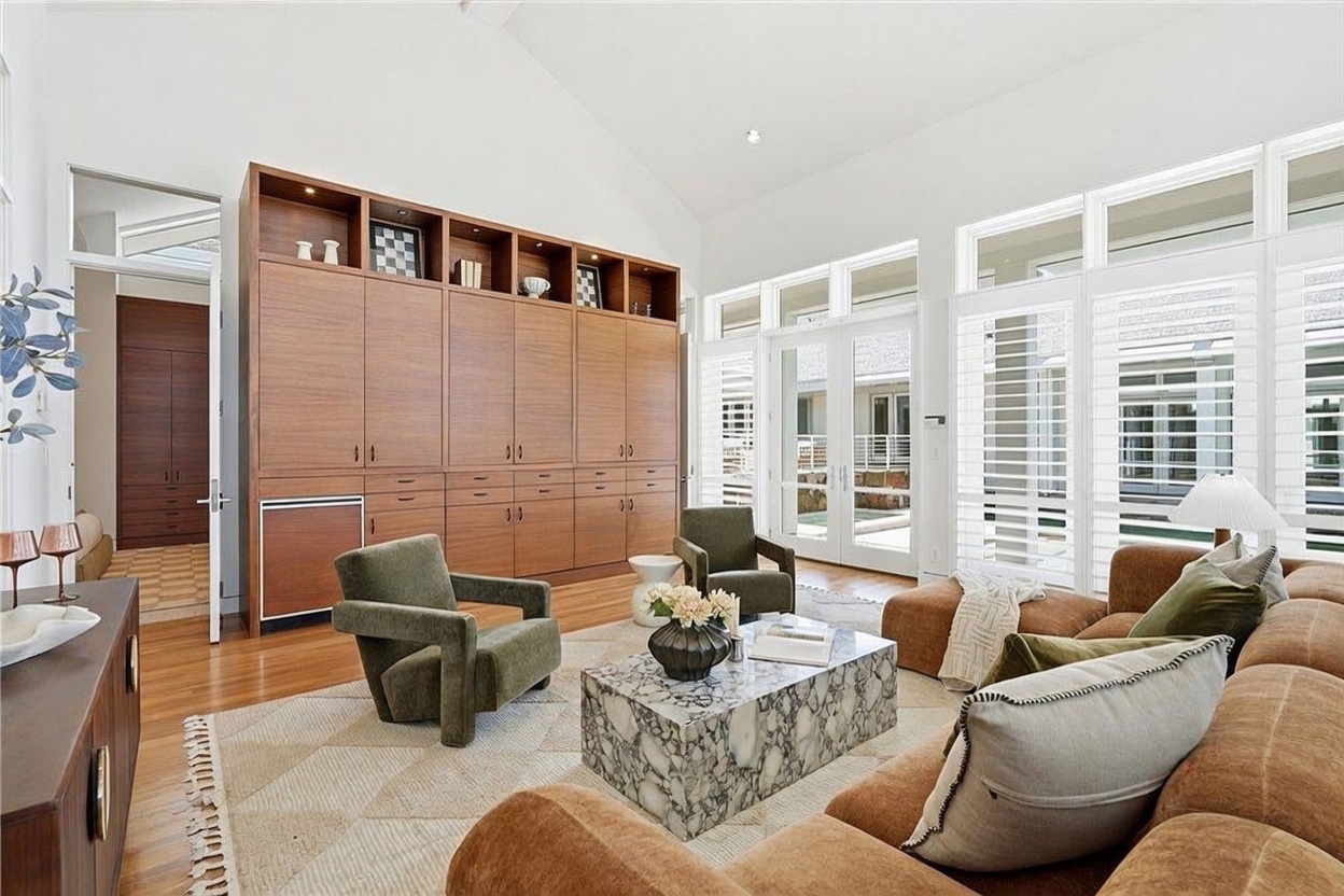 This spacious living room features high vaulted ceilings and a large wall of windows with white shutters that allow for abundant natural light. The room is anchored by a large, textured sofa and two olive-toned armchairs arranged around a stone-patterned coffee table. A prominent, custom-built wood cabinetry unit spans the wall, providing extensive storage and display space, while the light-toned wood flooring adds warmth to the bright, airy atmosphere.