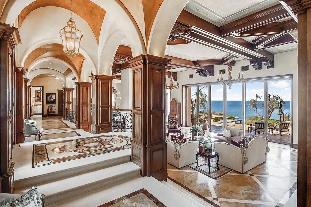 This expansive, Mediterranean-inspired living space features a grand arched hallway with intricate stone flooring and a sunken seating area that opens directly to an ocean view through large glass doors. The room is defined by heavy wood-beamed ceilings, ornate chandeliers, and rich wood columns that create a sense of architectural grandeur. The perspective is captured from the elevated hallway, looking down into the bright, airy living room which is furnished with plush, light-toned seating.