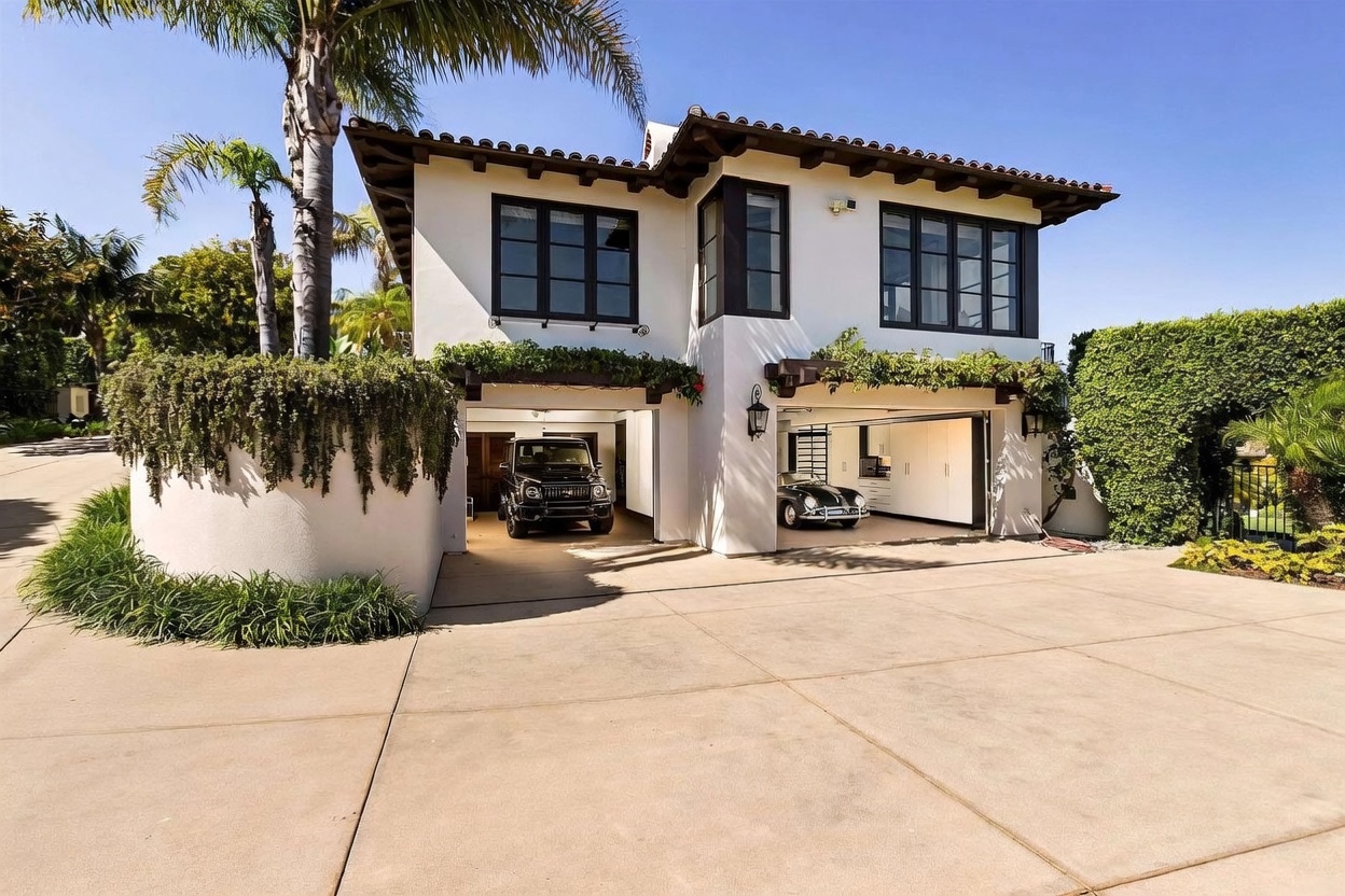 This Mediterranean-style home features a white stucco exterior with dark-framed windows and a tiled roof. The front of the house showcases a spacious two-car garage with an open layout, housing a black SUV and a vintage sports car. A large palm tree stands to the left, and the property is framed by lush greenery and a concrete driveway, creating a sophisticated and well-maintained curb appeal.